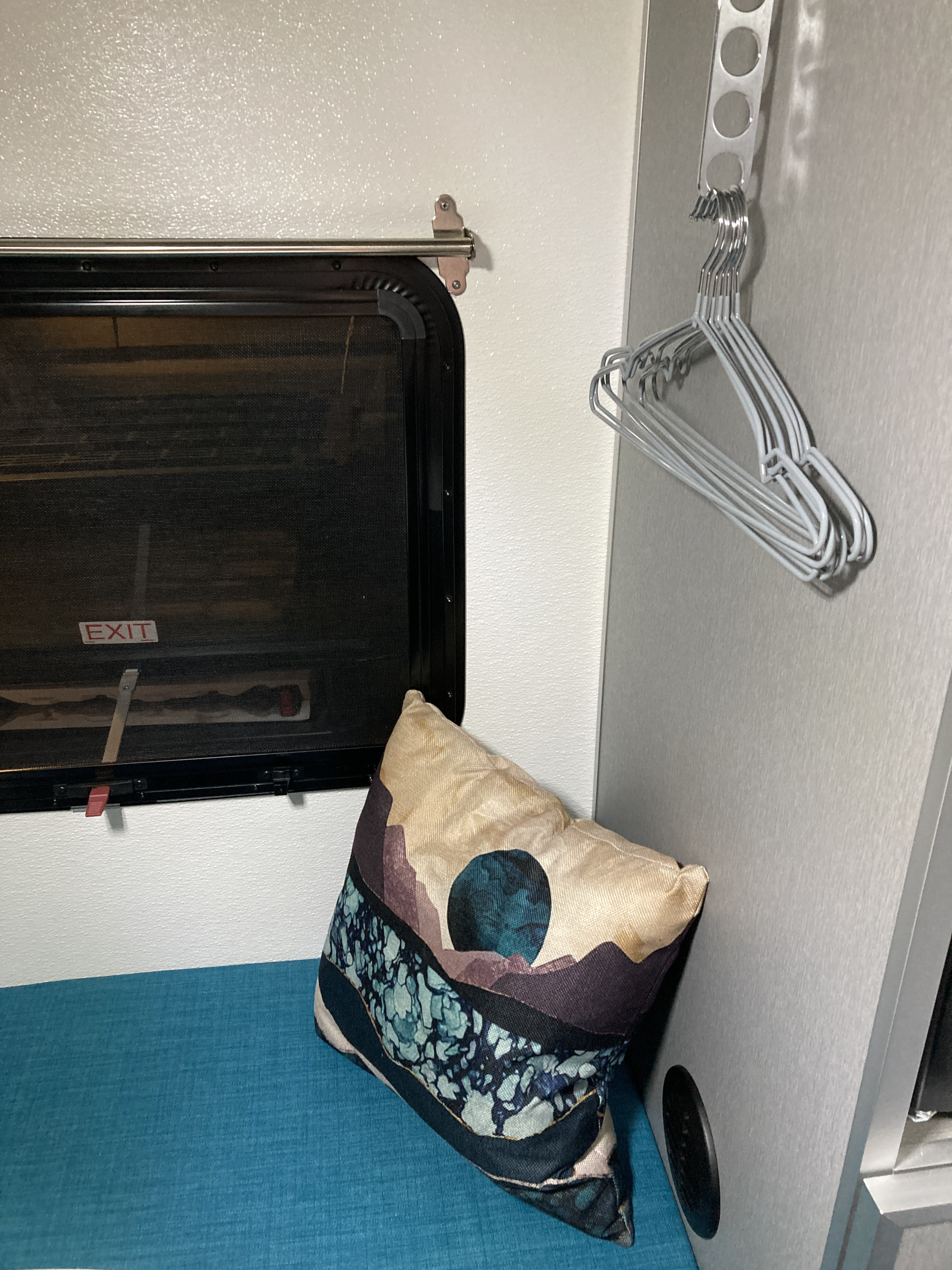 An RV rental's compact interior features an emergency exit window, a vibrant abstract pillow, and a practical clothes hanger rack.. Helio Helio Trailer 2023