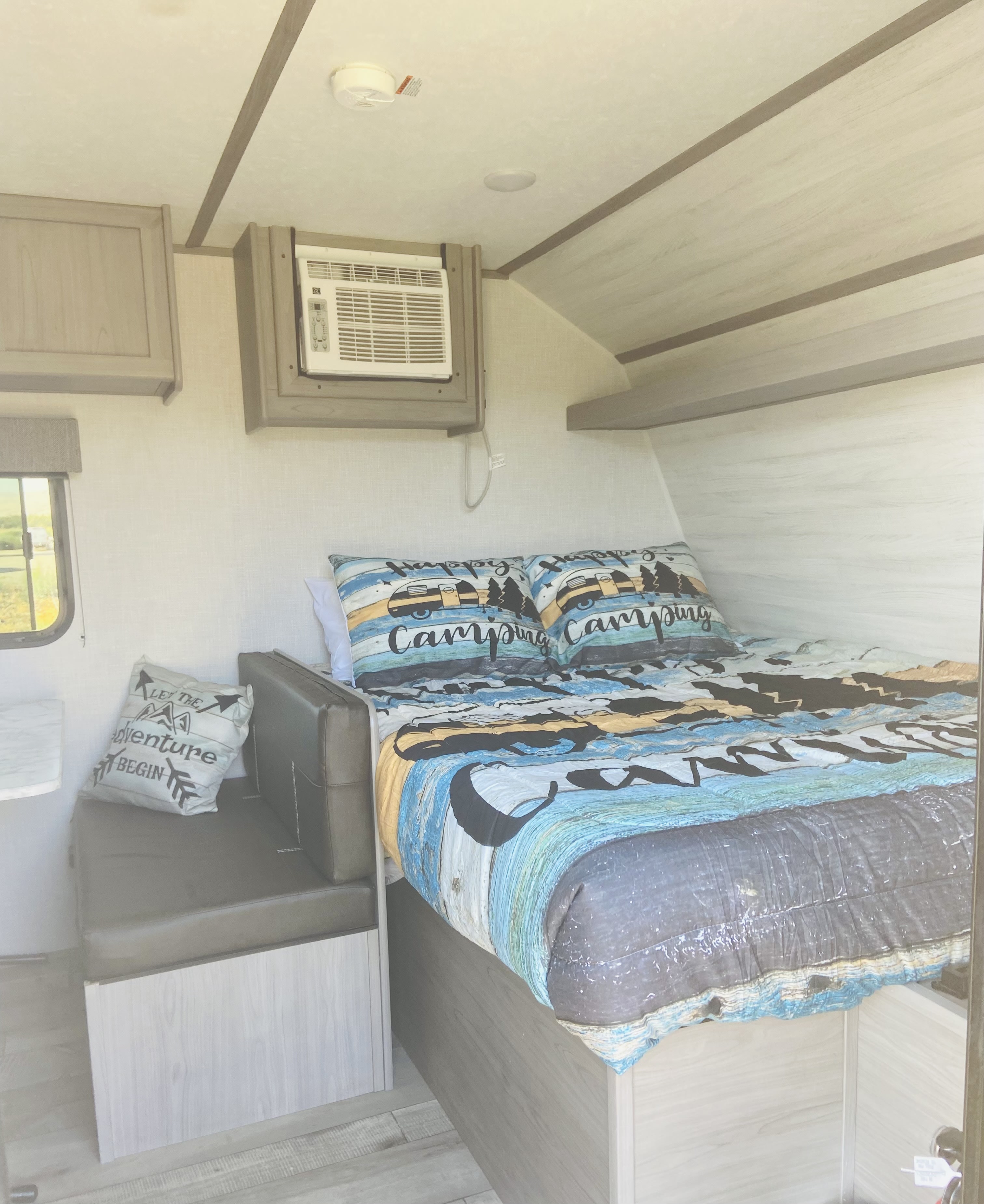 RV rental interior: light wood tones, a bed with colorful camping bedding, small dinette, and wall AC unit.. Dutchmen Coleman Lantern LT 2023