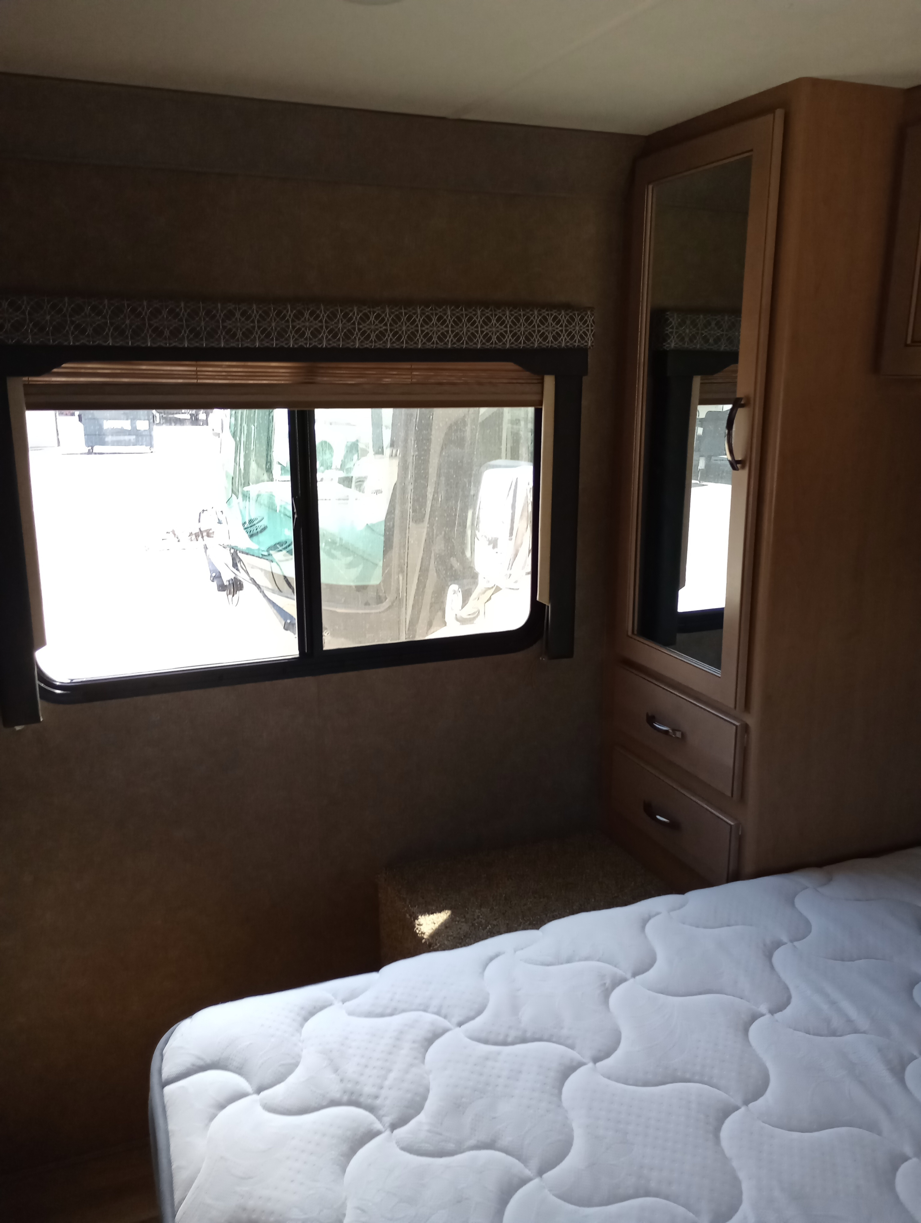 Cozy RV rental bedroom featuring a white quilted mattress, window, and wood wardrobe with mirror and drawers.. Thor Motor Coach Four Winds 2021
