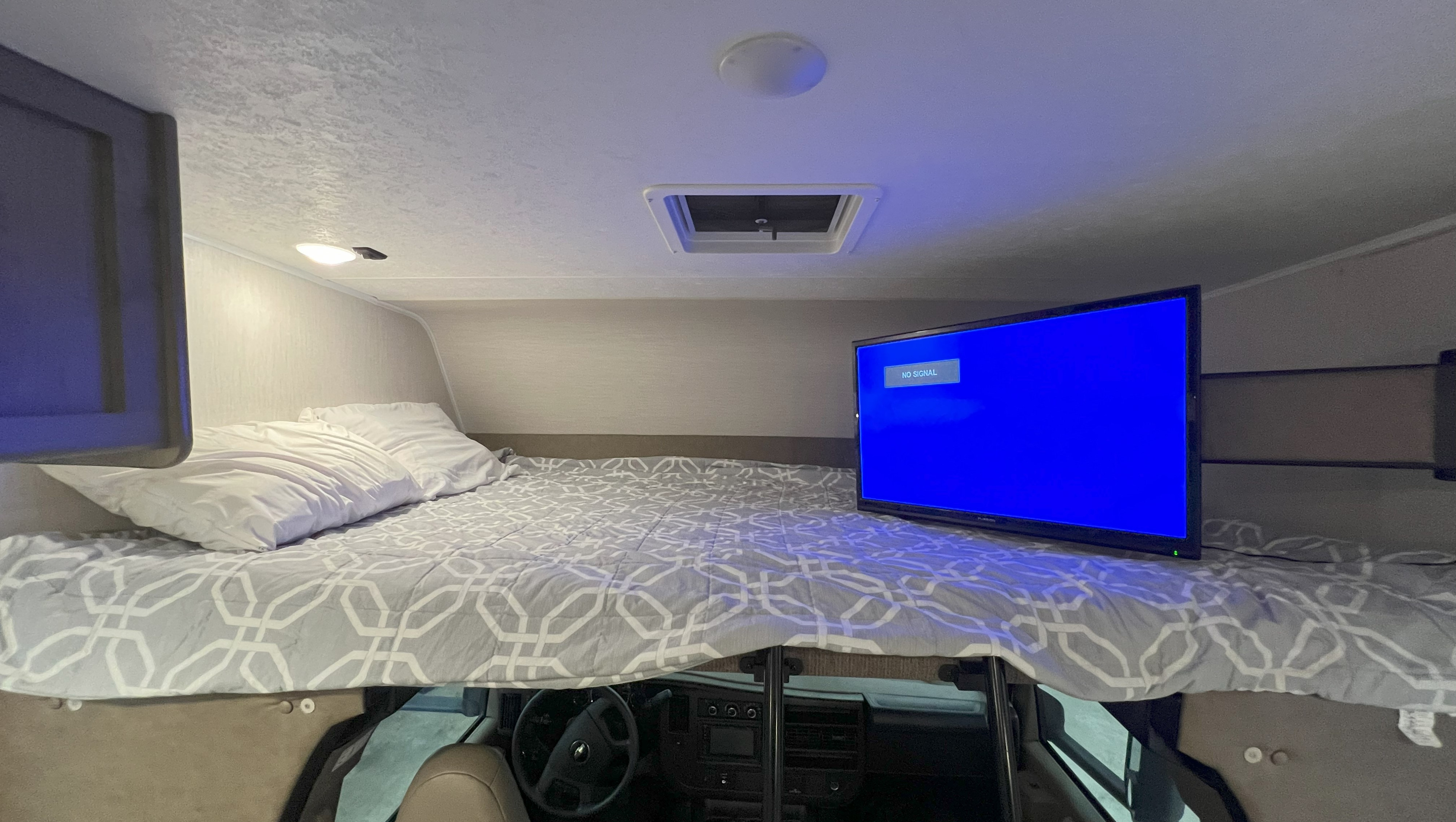 Cozy RV rental over-cab bunk bed with patterned comforter, pillows, TV showing 'NO SIGNAL', and cockpit view below.. Coachmen Freelander 2020
