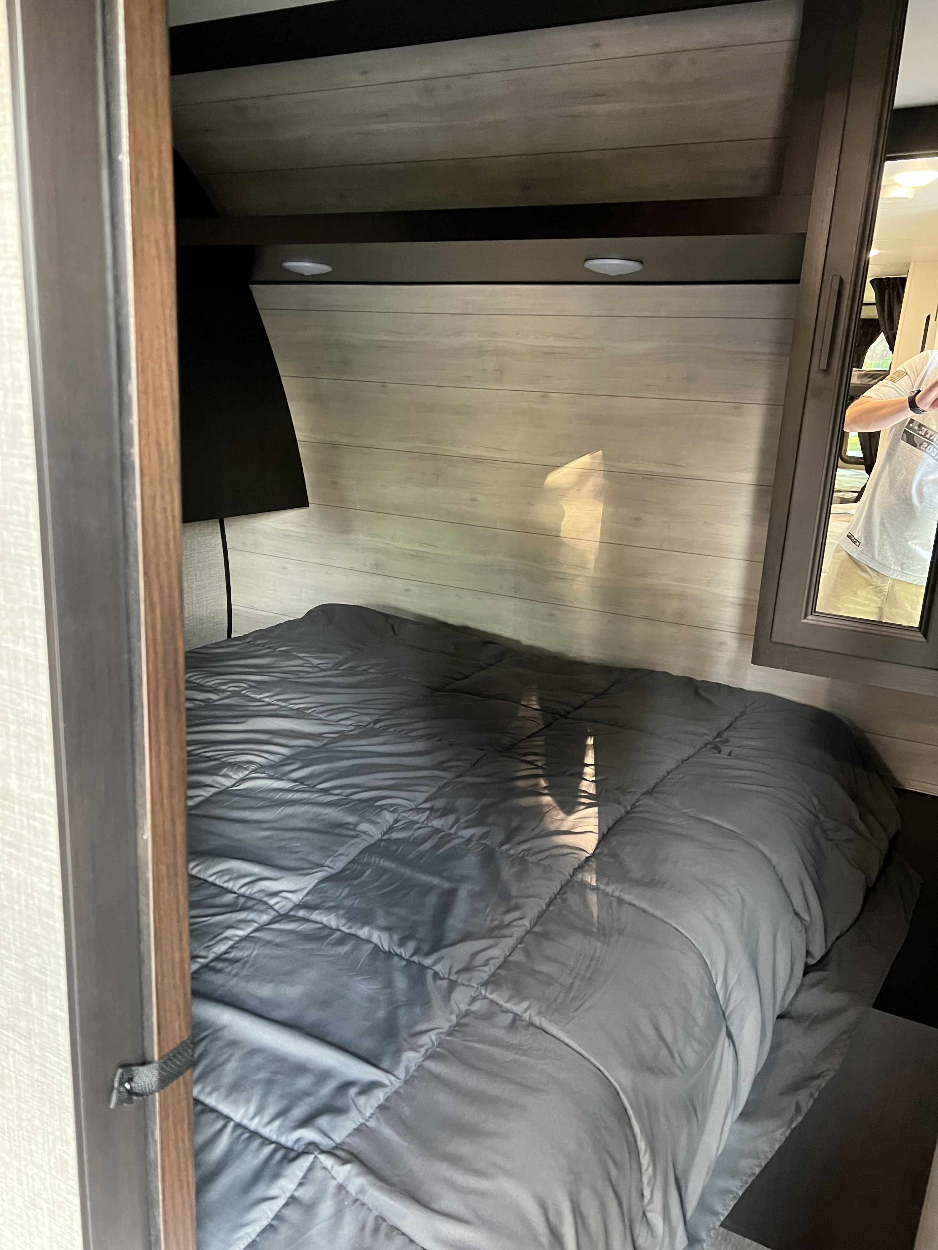 View into a comfy RV rental bedroom: grey bed, overhead storage, integrated lighting, and a mirror on the right.. Jayco Jay Flight 2023