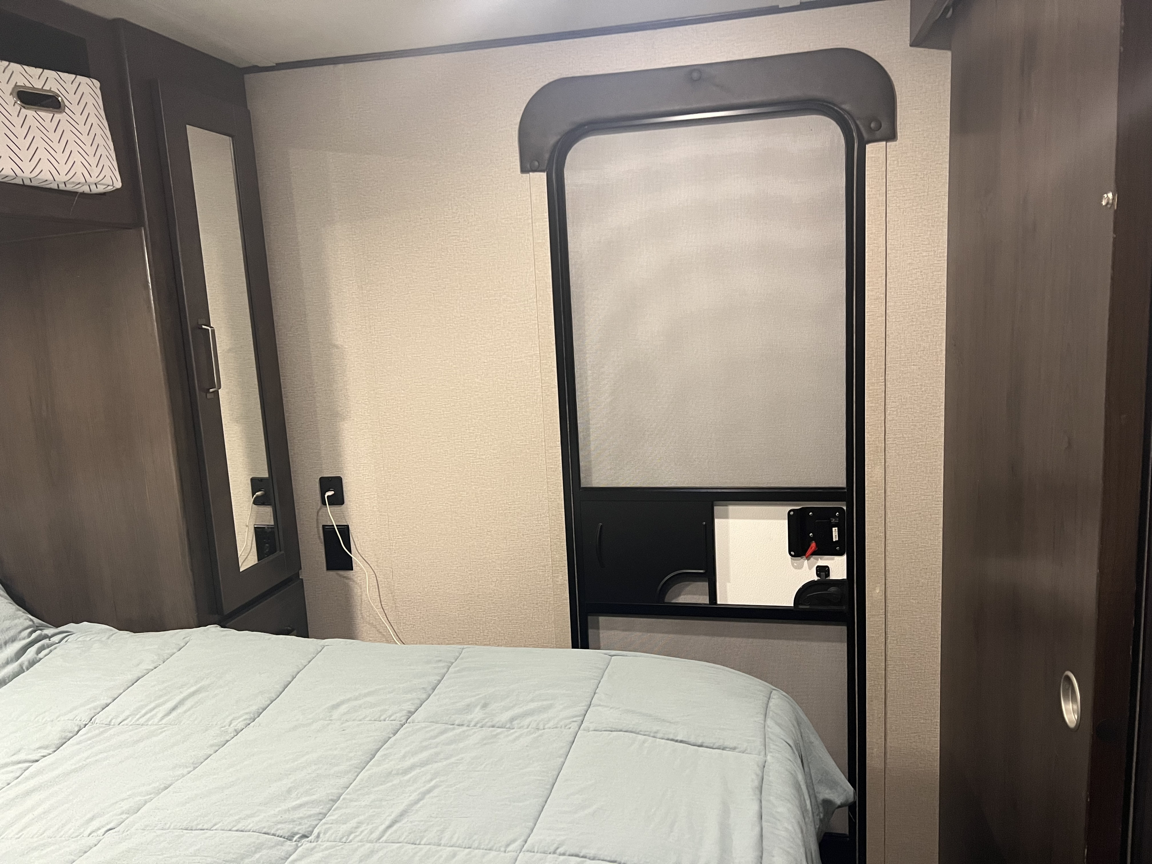 This RV rental bedroom features dark wood cabinetry, a light blue bed, and a prominent screen entry door, offering a compact, cozy space.. Grand Design Transcend Xplor 2022