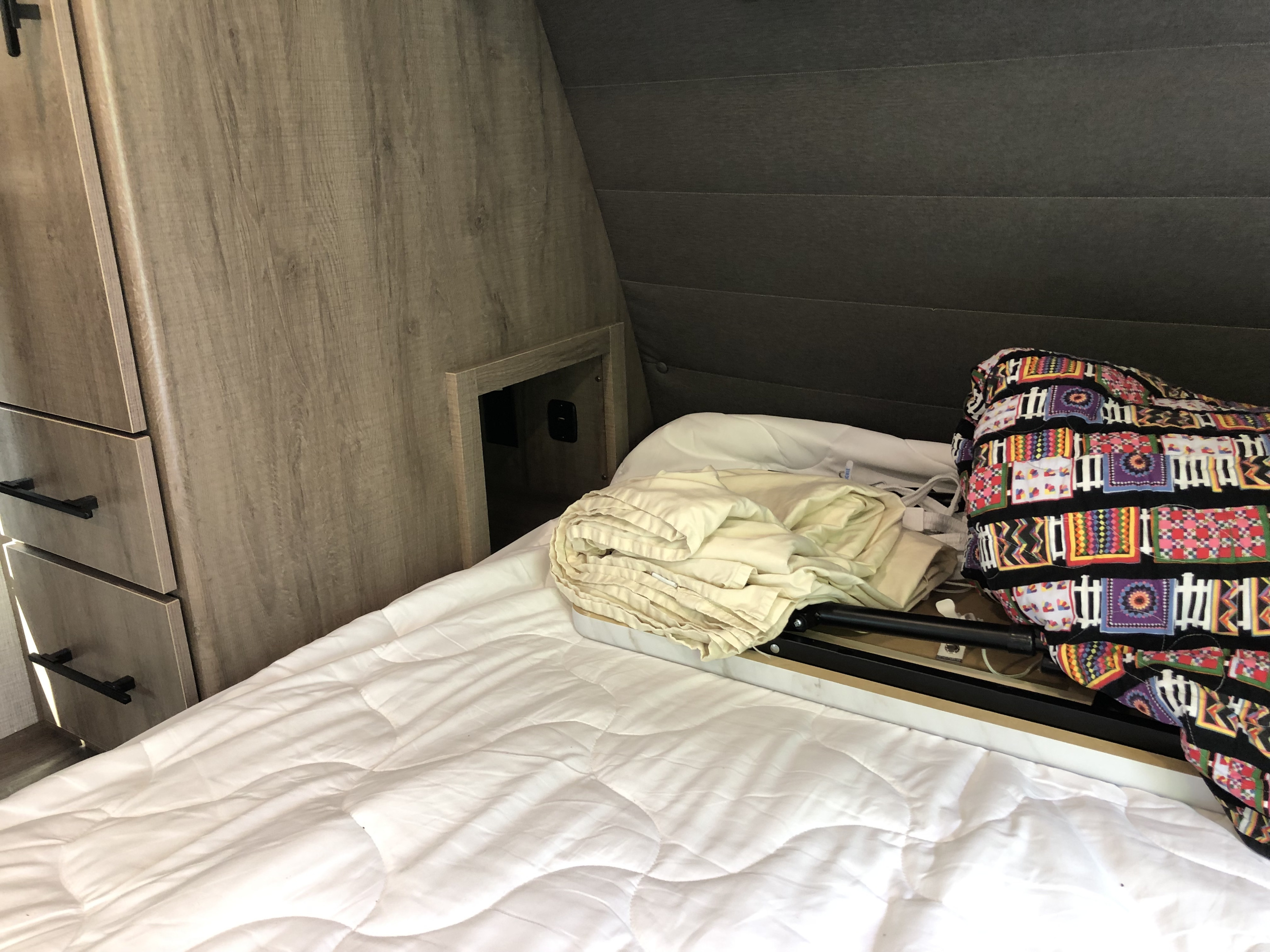 Cozy RV rental bedroom showing a white quilted bed, wood cabinets, and an open storage nook with power. Colorful patterned blanket and folded linens visible.. Grand Design Imagine 2023