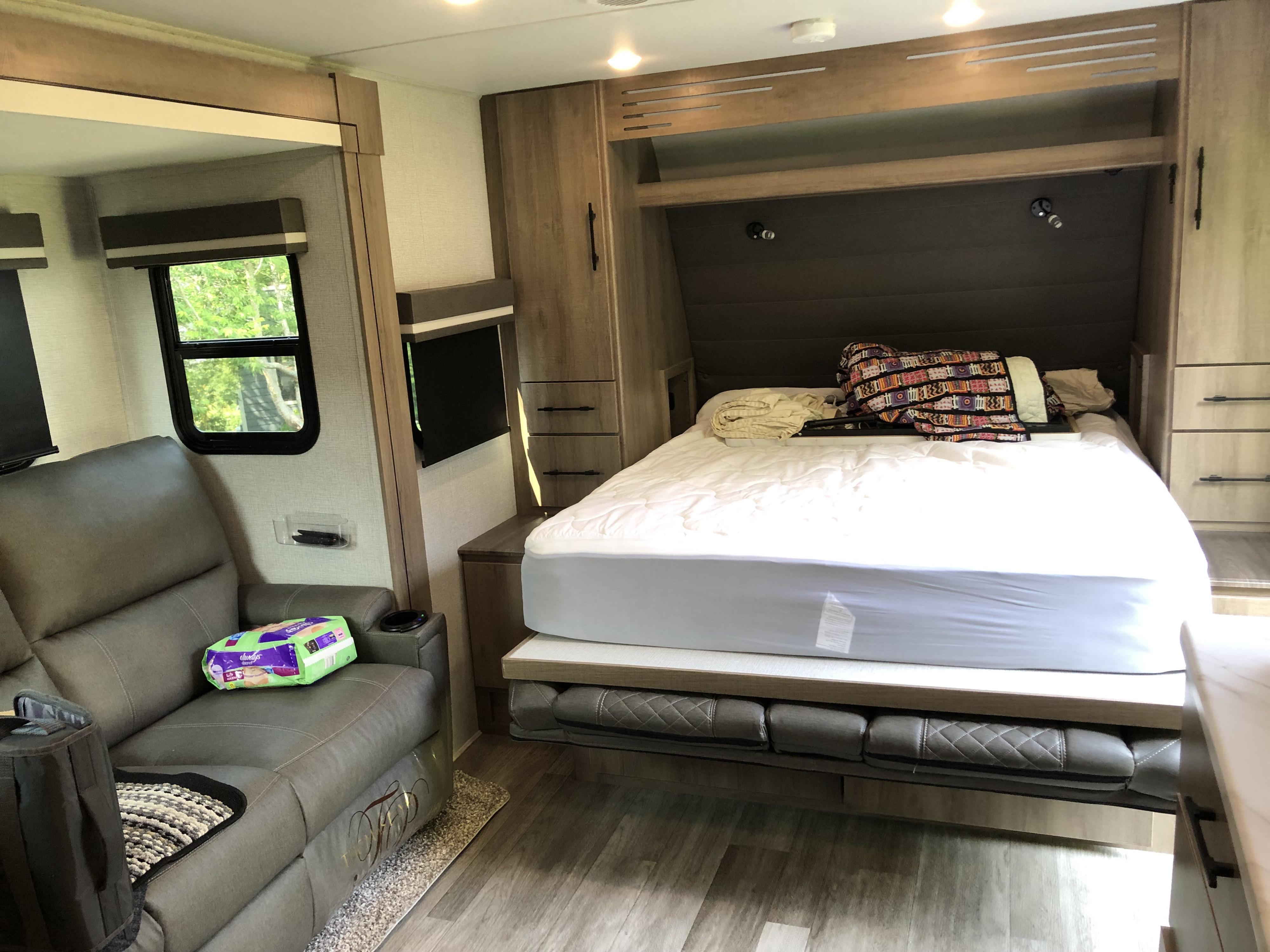 An RV rental interior showing a grey reclining sofa and a space-saving Murphy bed setup with a mattress.. Grand Design Imagine 2023