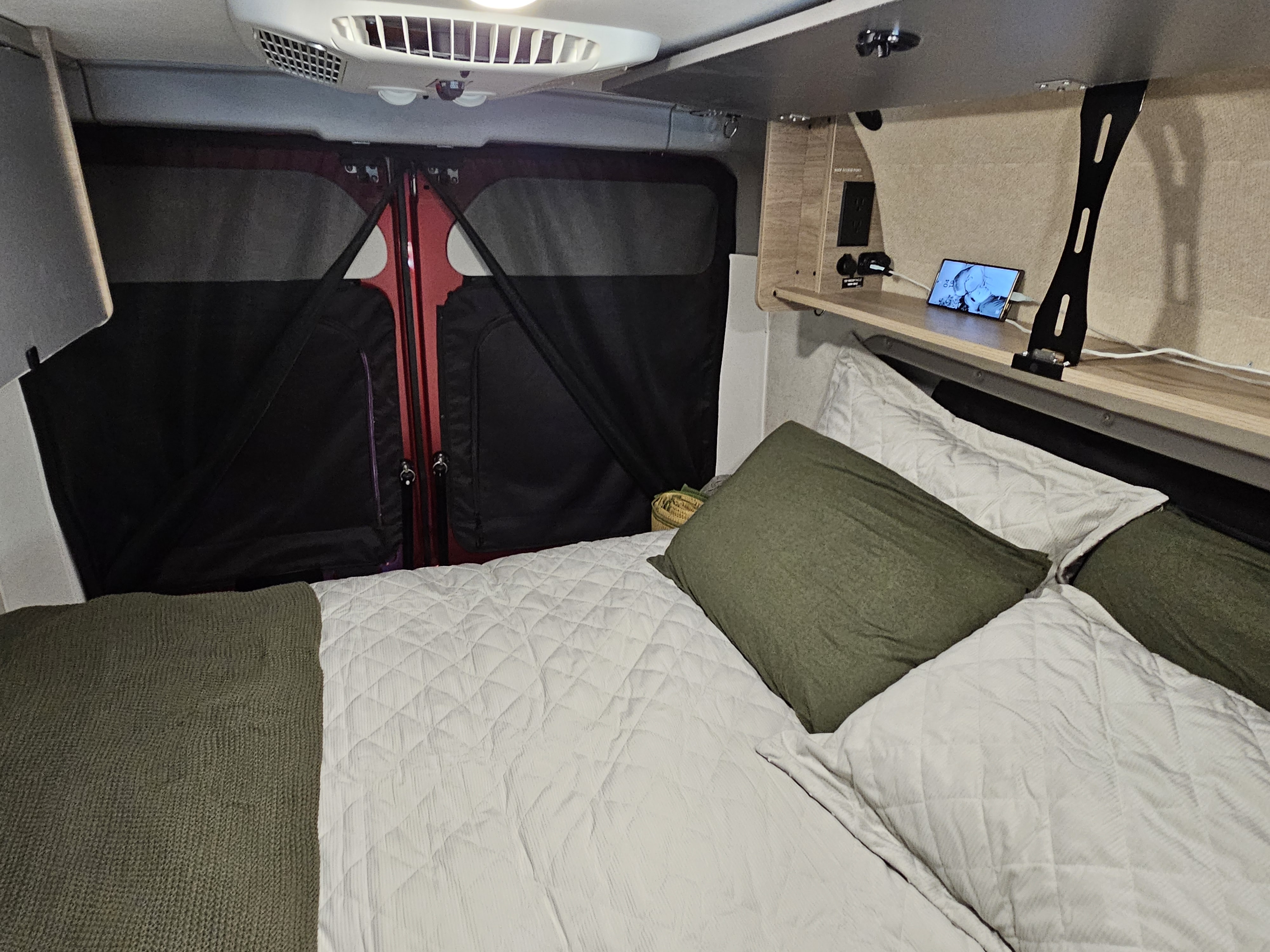 Cozy RV rental interior with a bed, charging station, and privacy screens covering the red rear van doors.. Winnebago Solis 2023