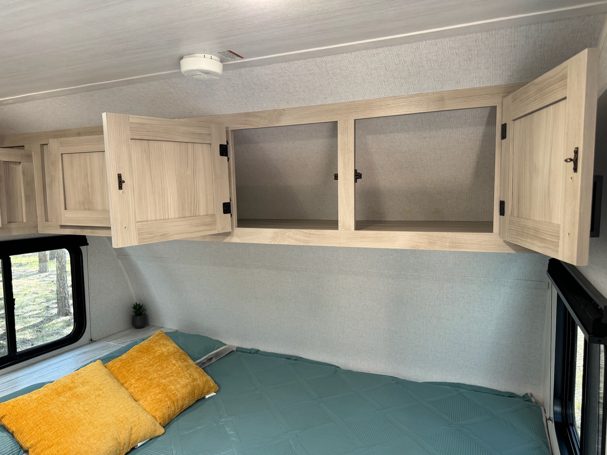 This RV rental bedroom features open overhead cabinets, a bed with teal bedding & yellow pillows, and a window view.. East to West Della Terra 181 BHSLE 2024