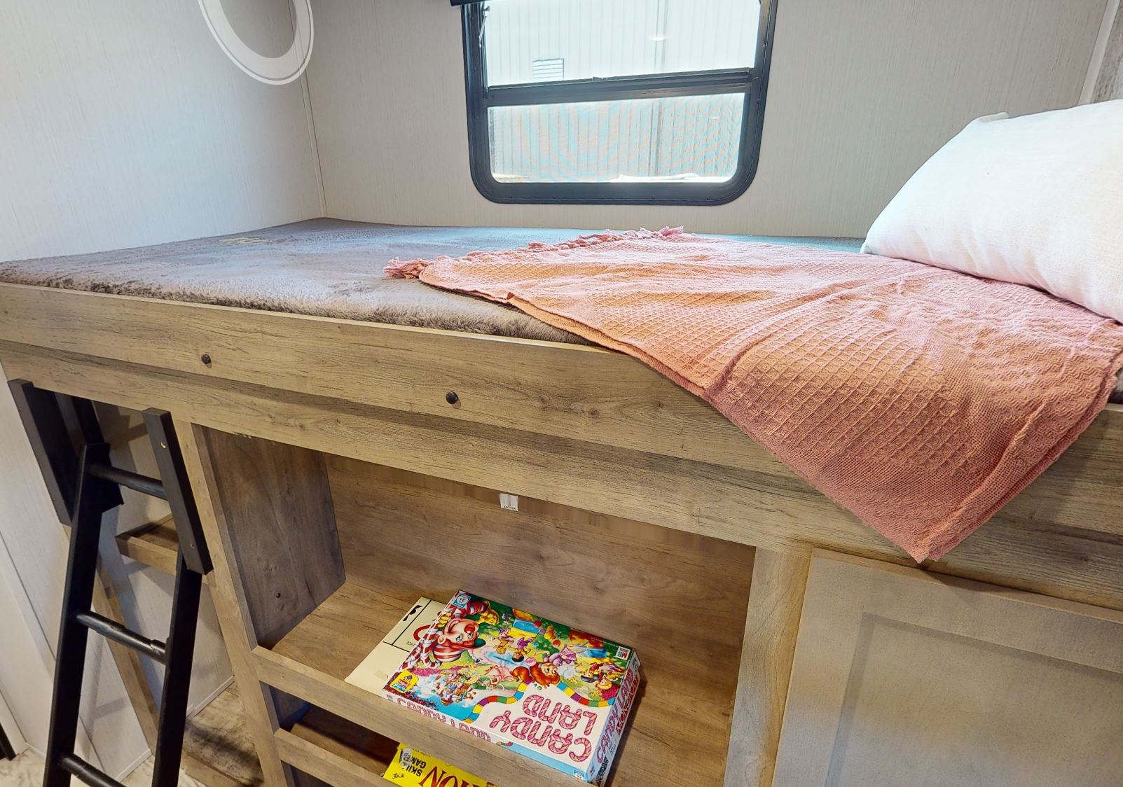 Cozy RV rental bunk bed with pink blanket, ladder, and under-bunk storage holding a Candy Land game.. Forest River Puma 2023