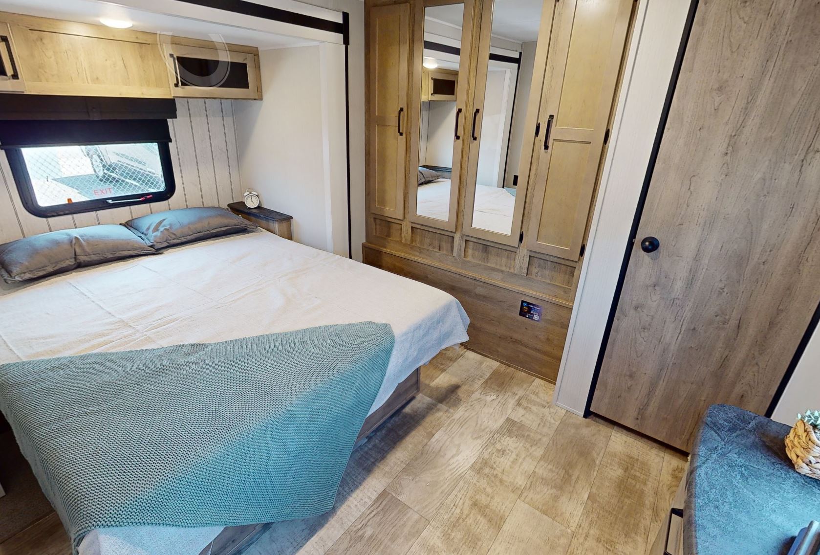 Spacious RV rental bedroom: cozy bed with blue throw, overhead cabinets, window, mirrored wardrobe, and wood flooring.. Forest River Puma 2023
