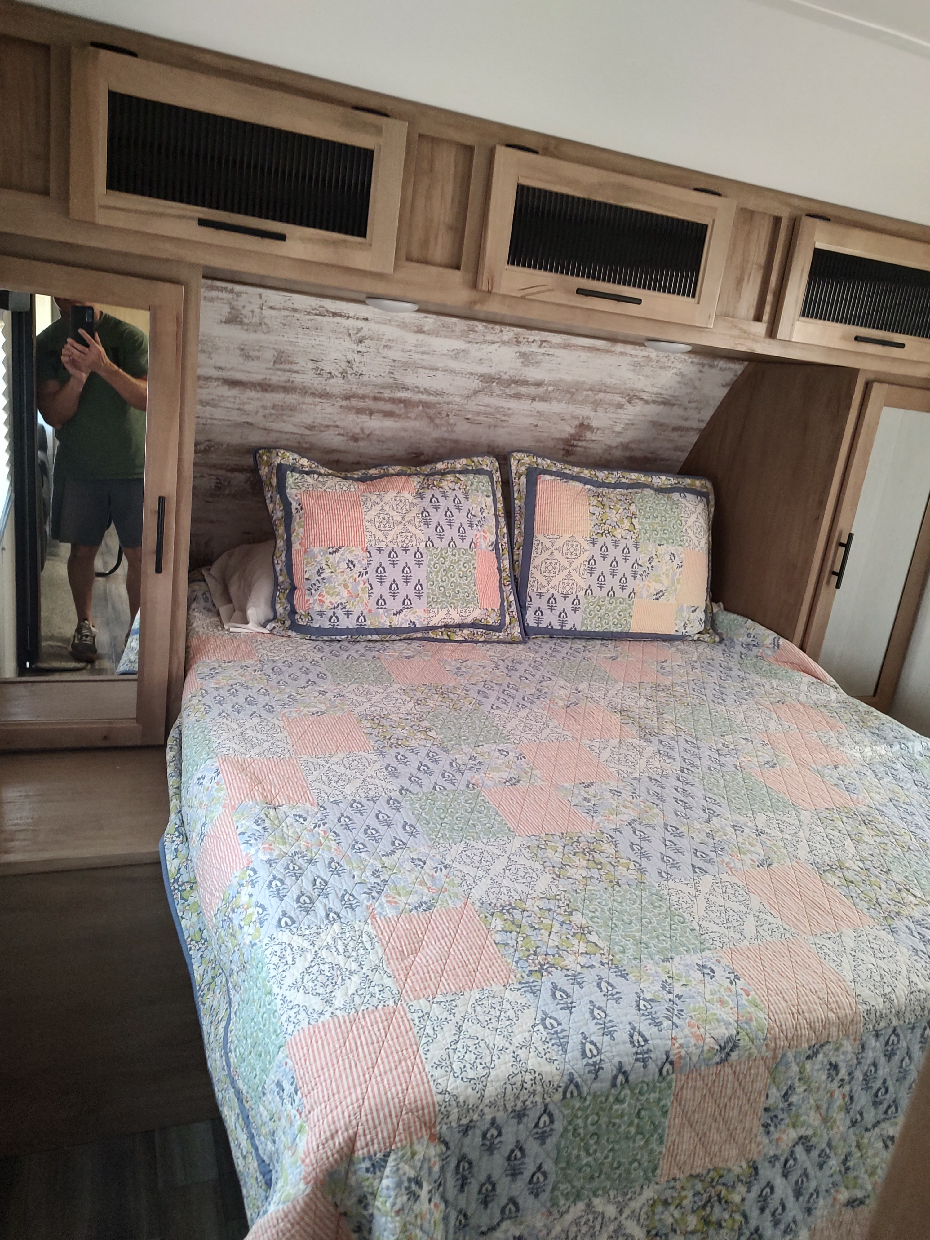 Cozy RV rental bedroom featuring a colorful patchwork quilt, overhead cabinets, and a mirror reflecting the photographer.. Forest River Cherokee Alpha Wolf 2023