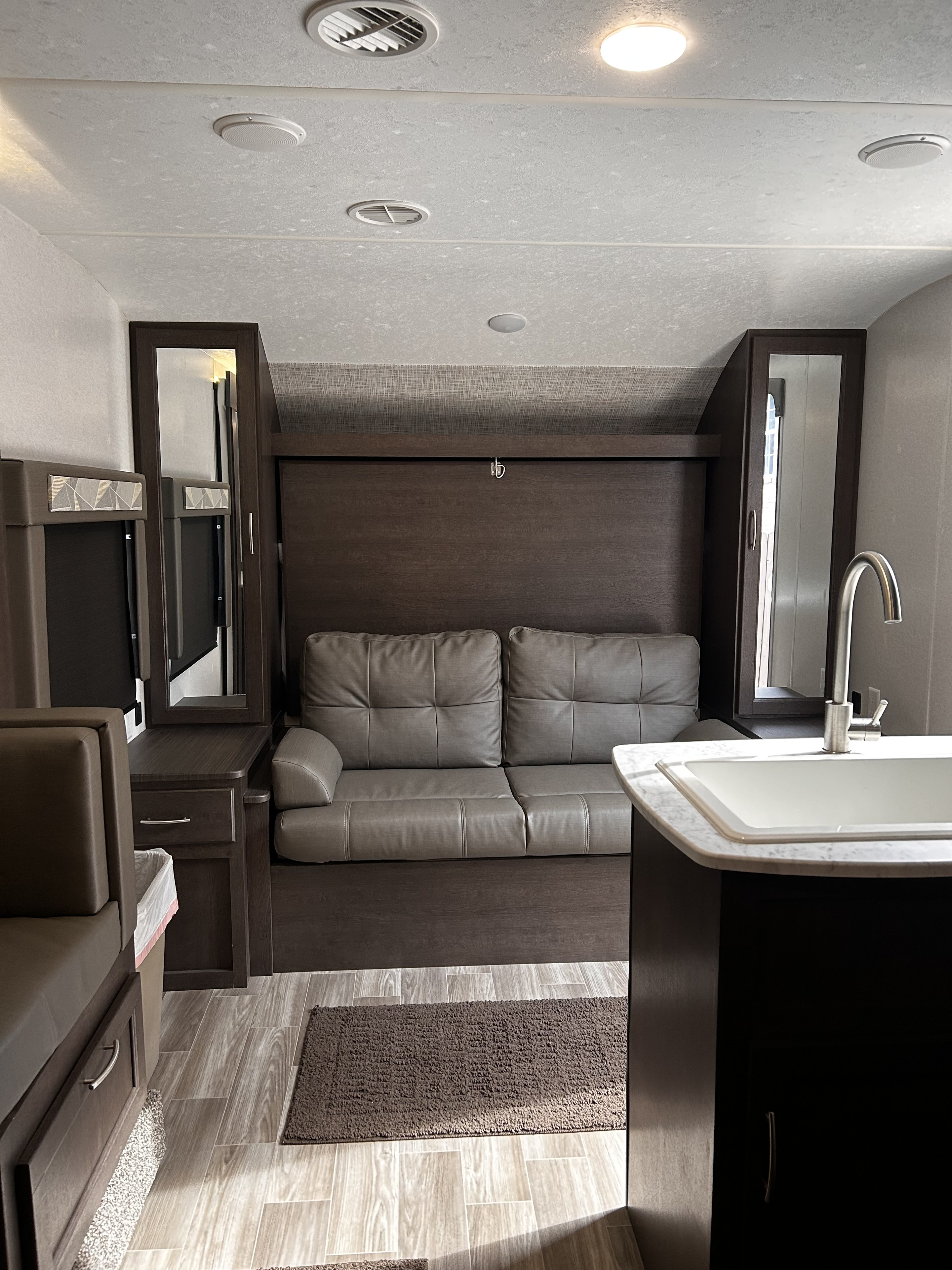 View of an RV rental interior featuring a gray sofa, dark wood cabinetry, kitchen sink, and wood-look flooring.. Forest River Wildwood Cruise Lite 2019