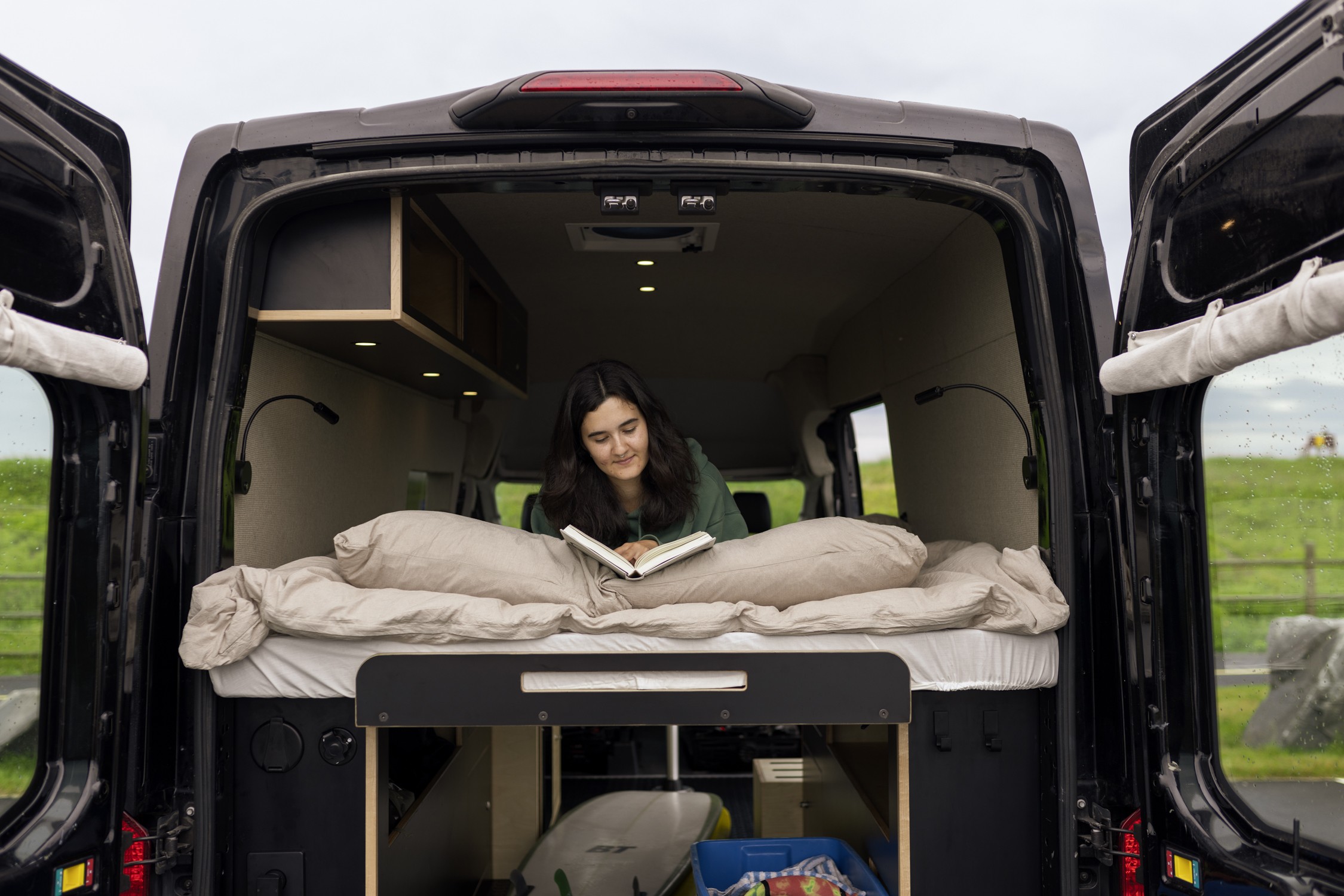 A young woman reads on a bed inside an RV rental, showcasing its spacious interior, built-in storage, and a surfboard.. Ford Transit 2020
