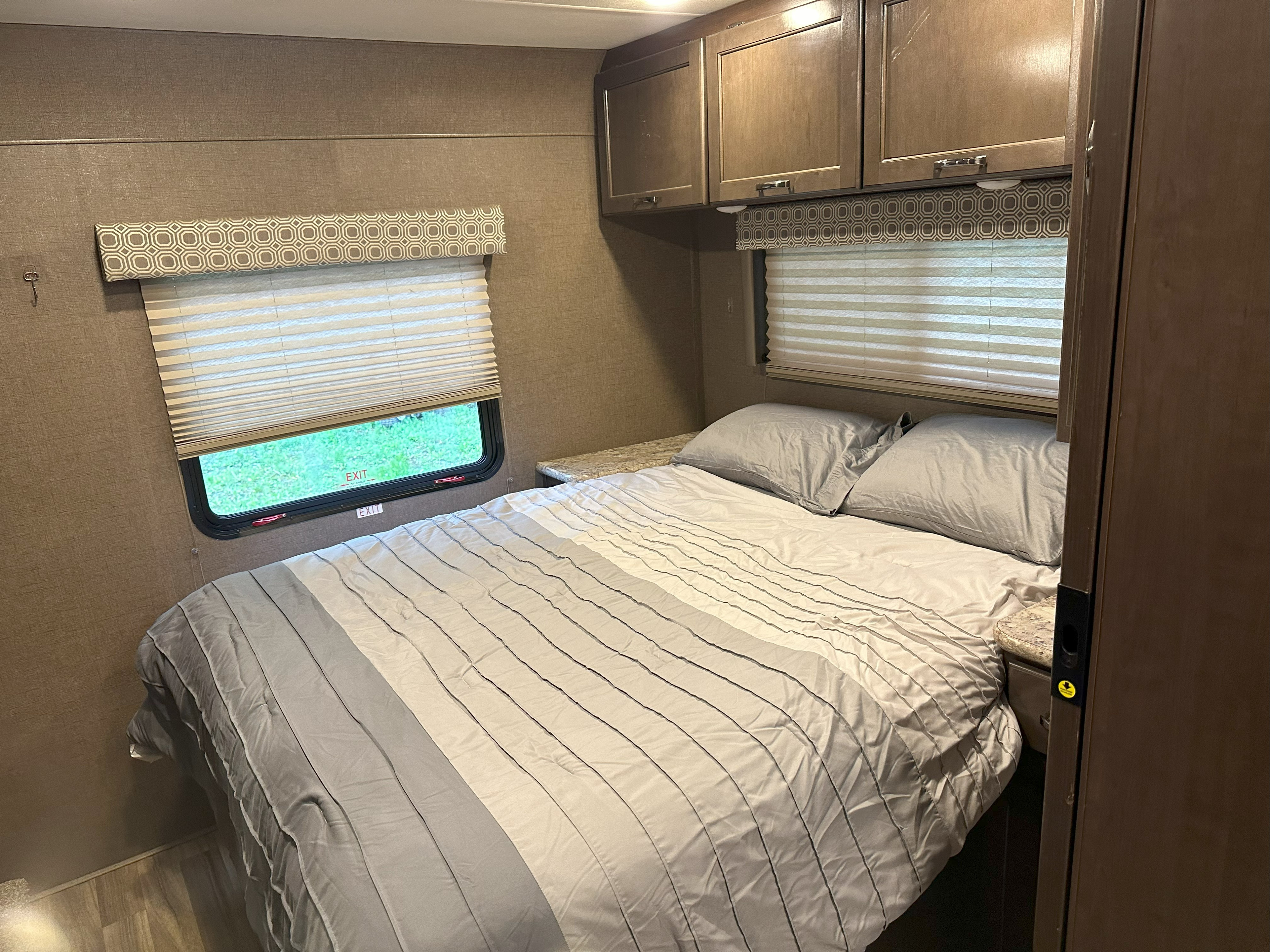 RV rental bedroom: a cozy bed with gray bedding, overhead cabinets, and a window with an "EXIT" sign in this compact space.. Thor Four Winds 2019