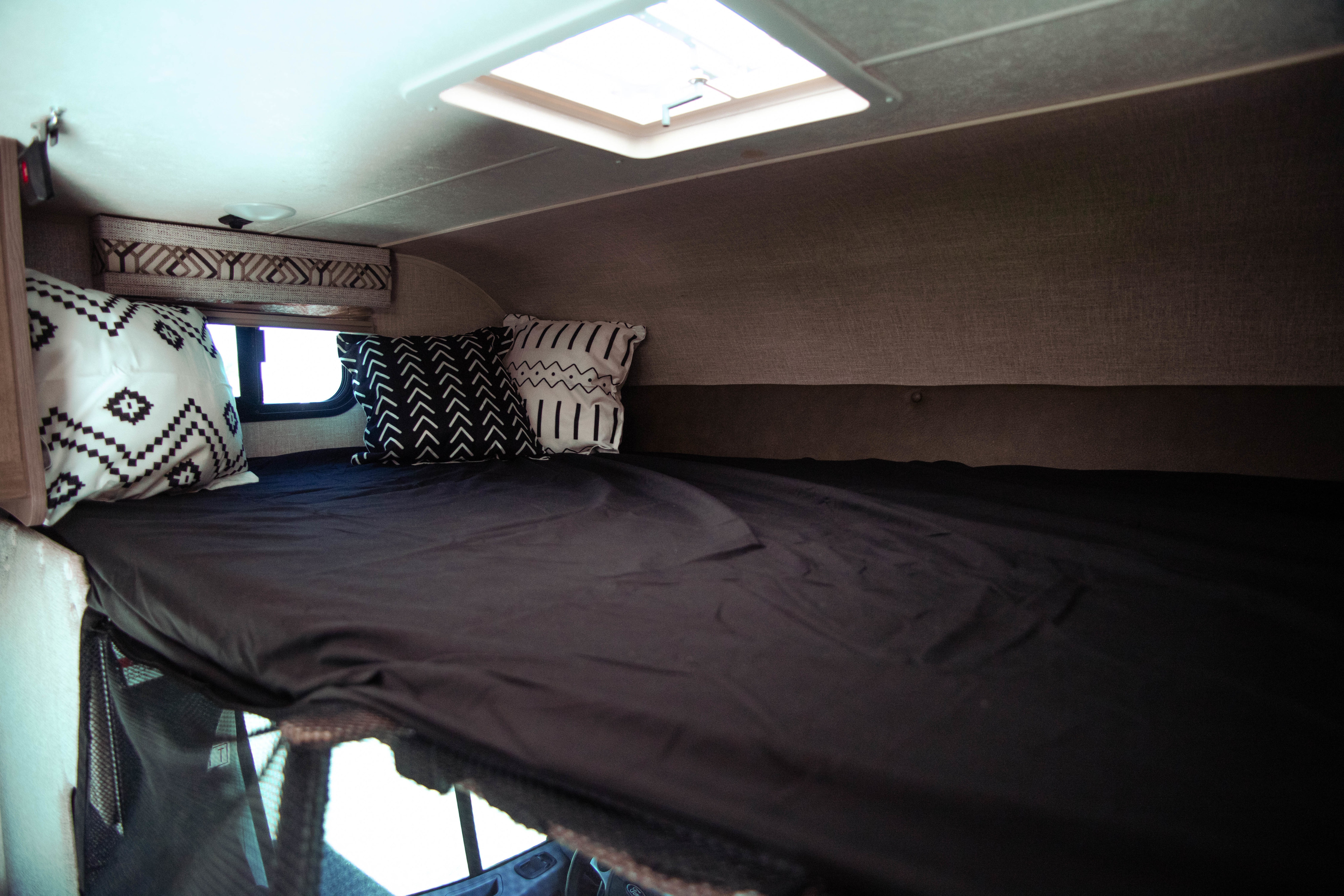 An RV rental's cozy cab-over bed area, featuring black sheets, patterned pillows, a window, and a ceiling skylight.. Coachmen Orion 2020