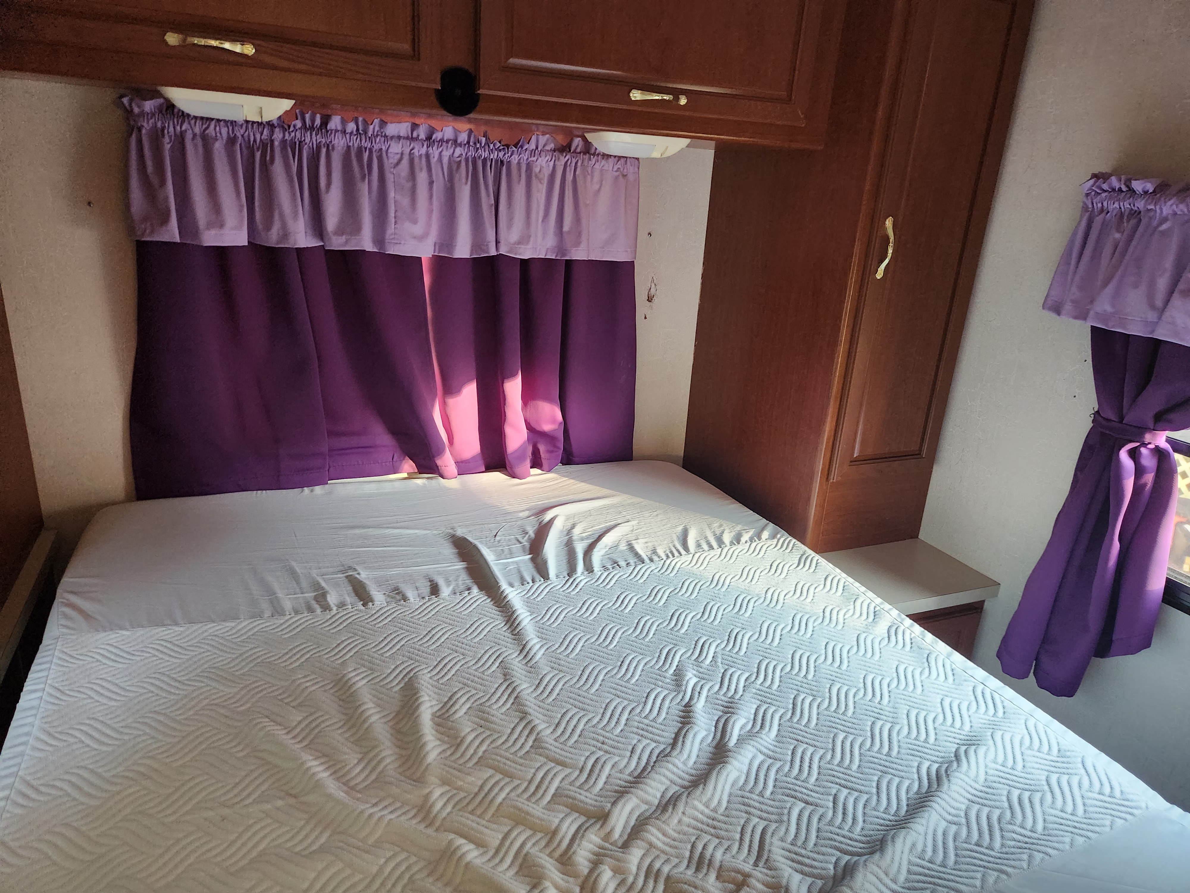 An RV rental bedroom featuring a bed with a white textured comforter, purple curtains, and built-in wooden cabinets.. Coachmen Mirada 2002