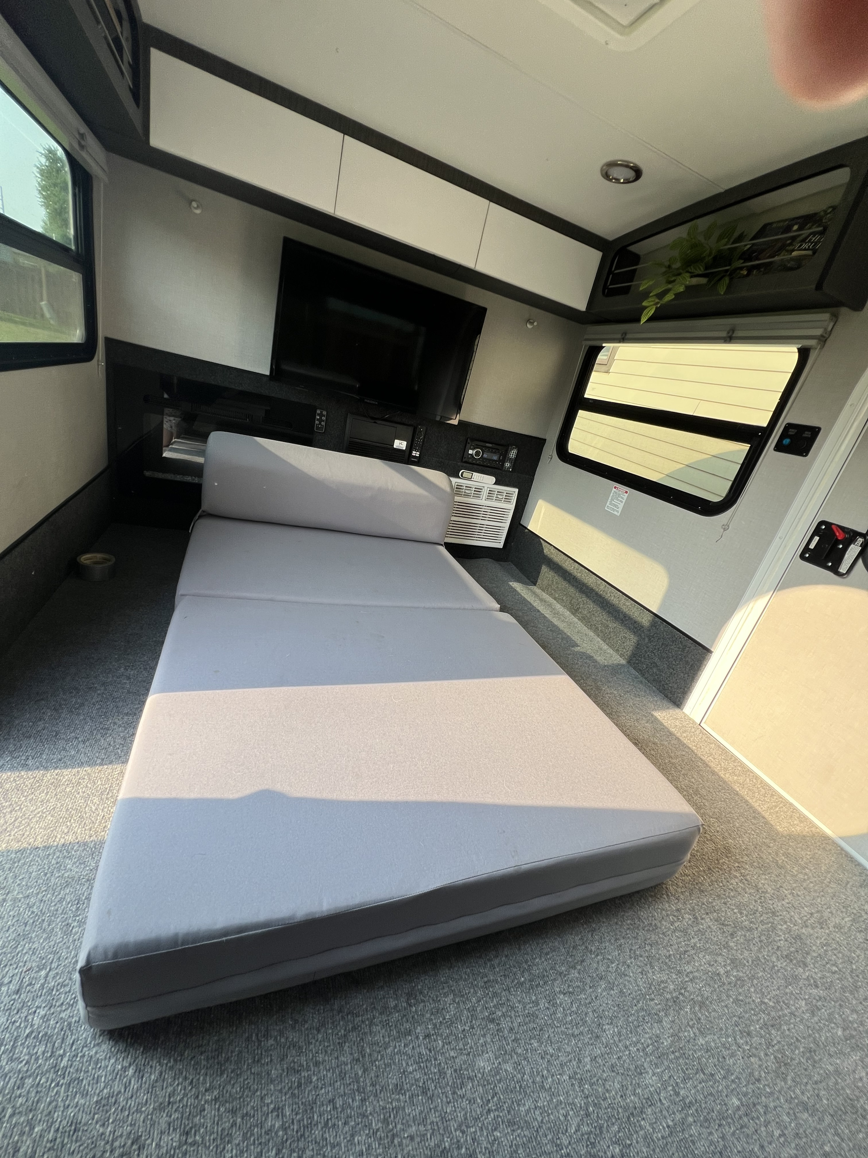 Inside a modern RV rental: a gray tri-fold mattress, TV, AC, overhead cabinets, and windows with blinds.. InTech RV Luna 2022