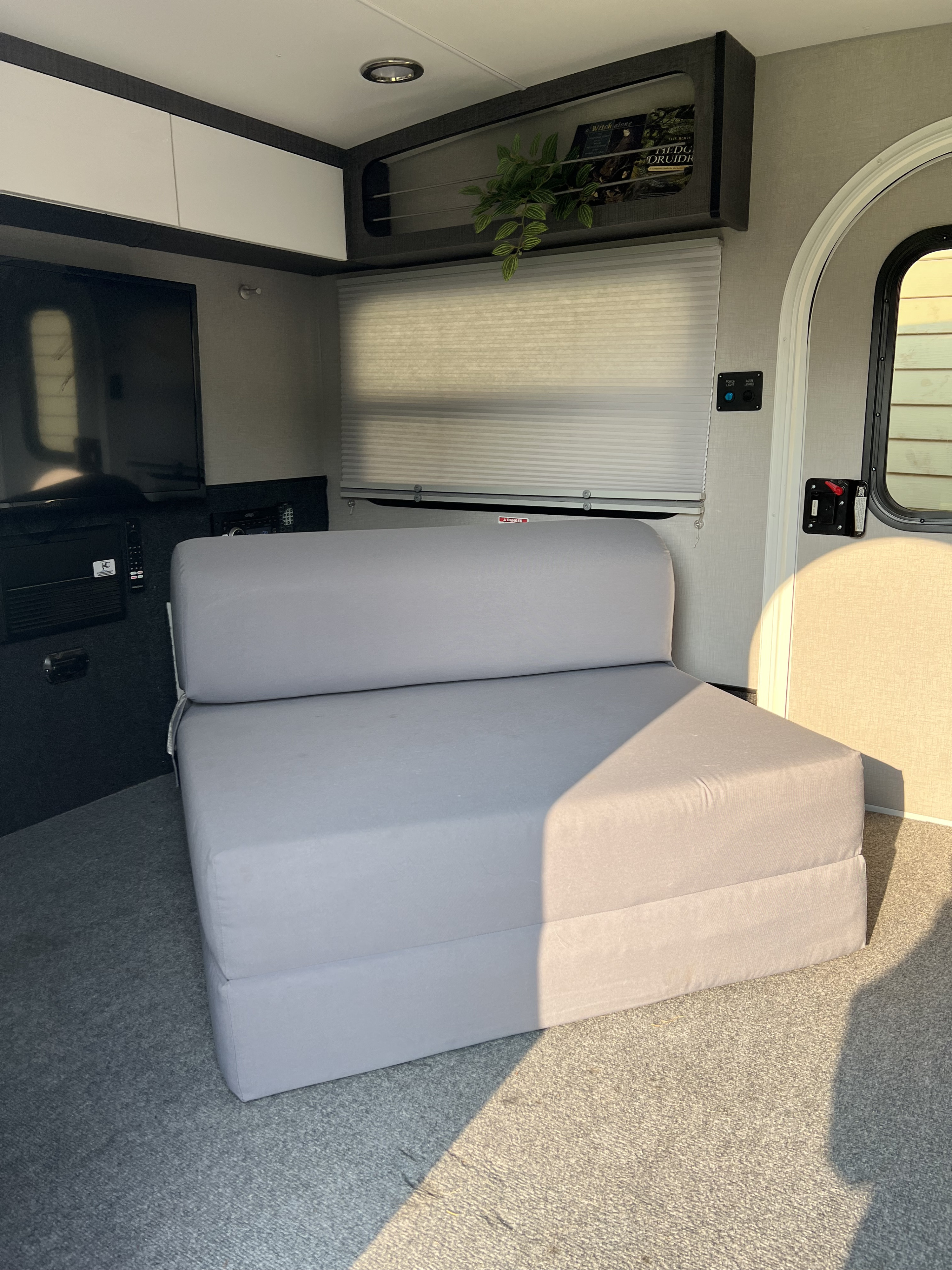 Inside this rv rental, a light gray foldable sofa sits on a carpeted floor. Overhead cabinets, a TV, and a window with blinds are visible.. InTech RV Luna 2022