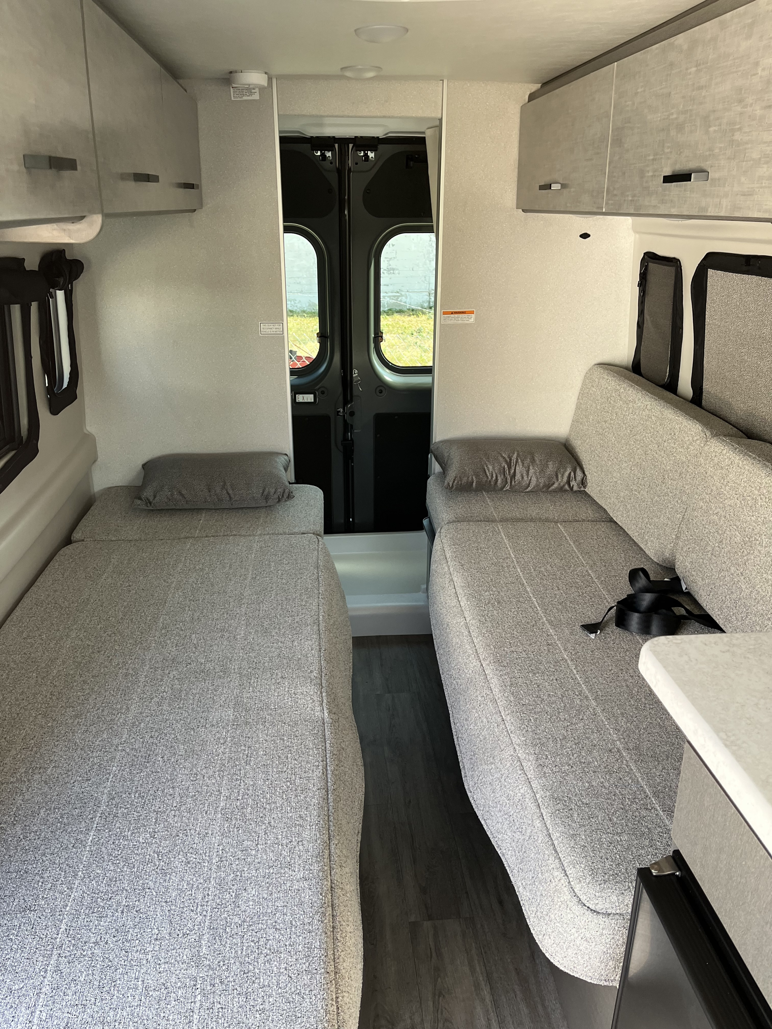 Clean rv rental interior shows two grey beds/benches, central aisle, overhead storage, and rear doors. Compact travel ready.. Thor Twist 2025
