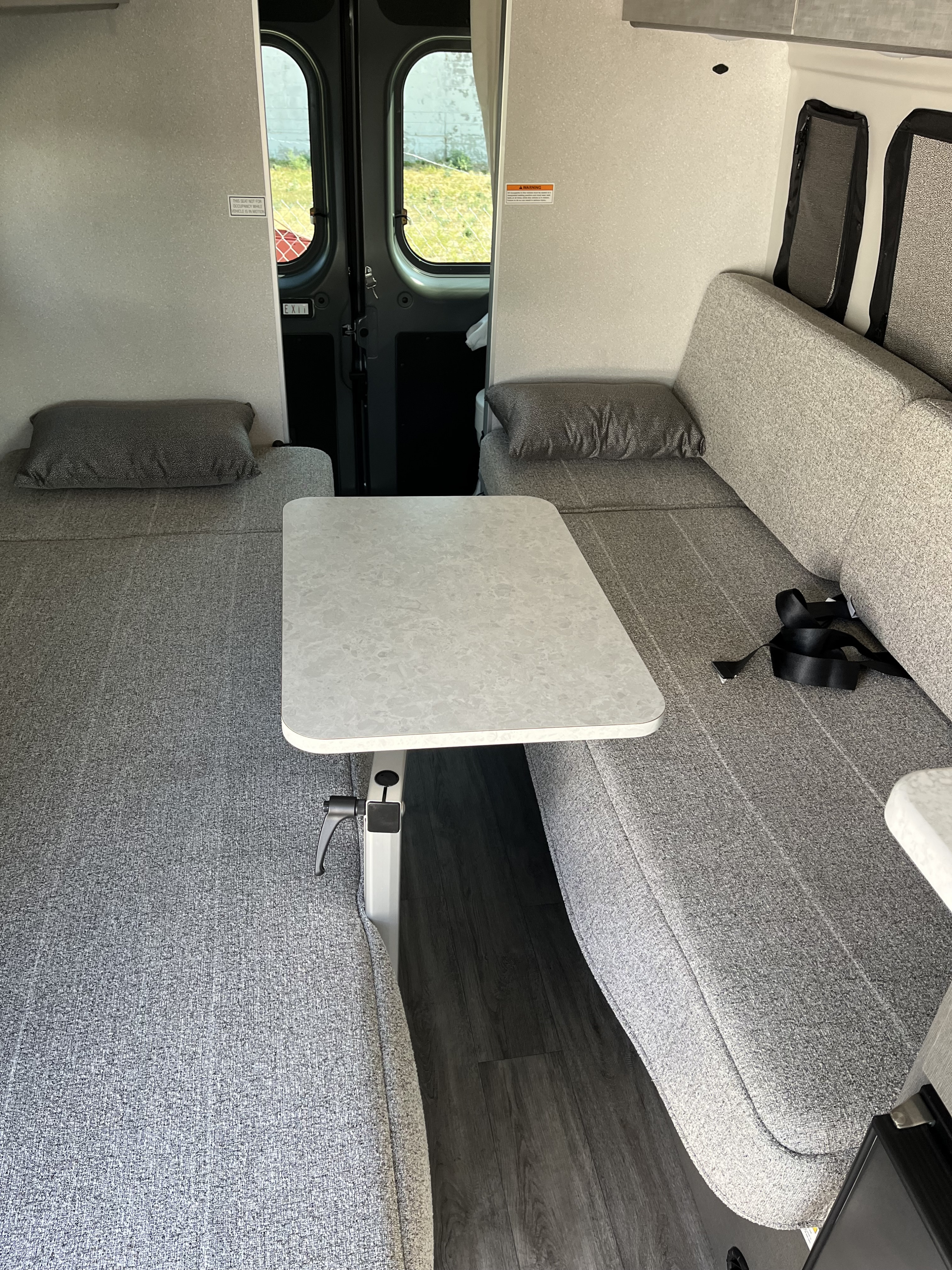 Cozy RV rental interior with grey benches converting to beds, a central table, and rear exit doors visible.. Thor Twist 2025