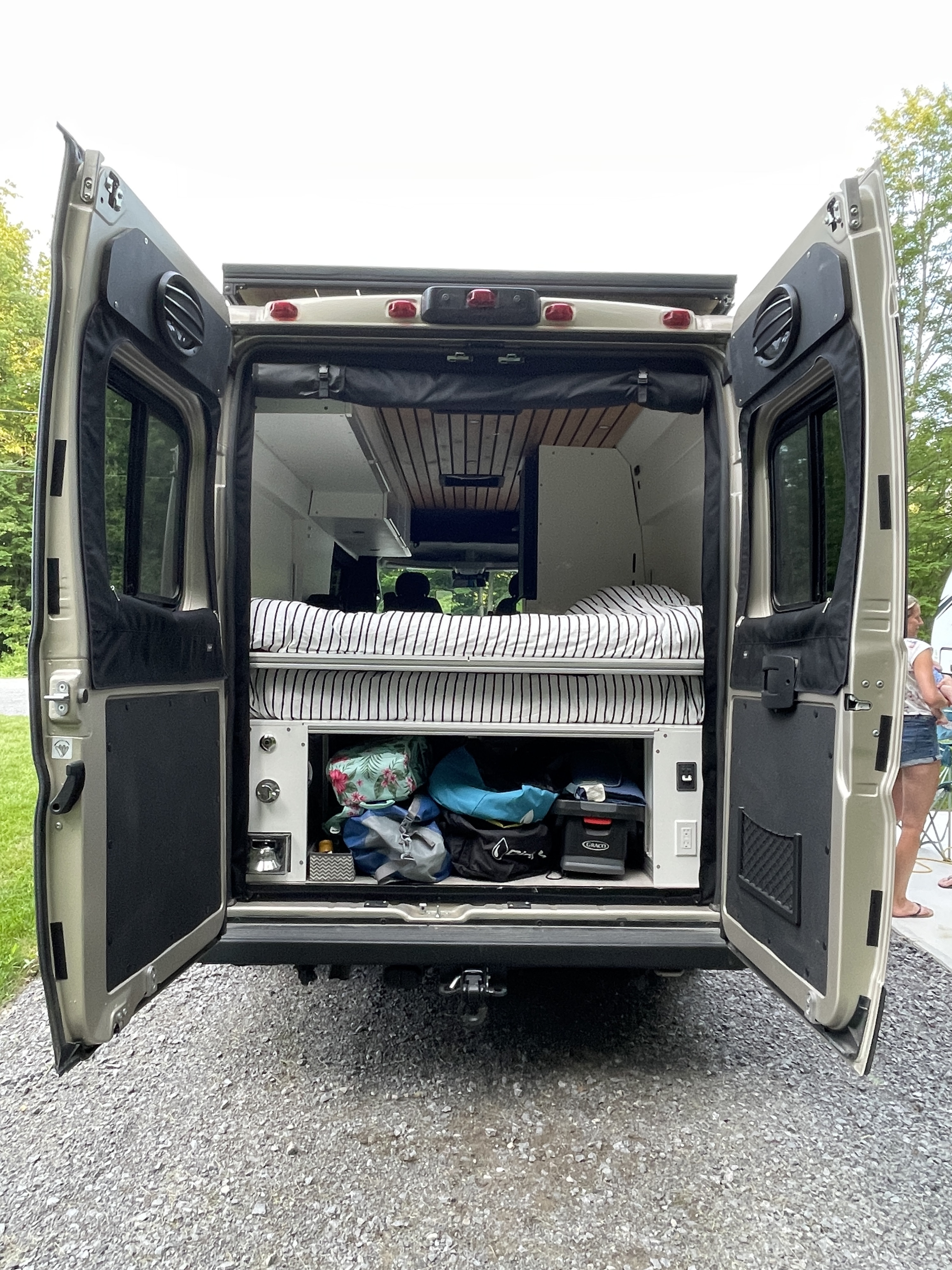 Peek into a cozy rv rental camper van! Rear doors open, revealing a striped bed & organized under-bed storage.. Ram Promaster 3500 2023