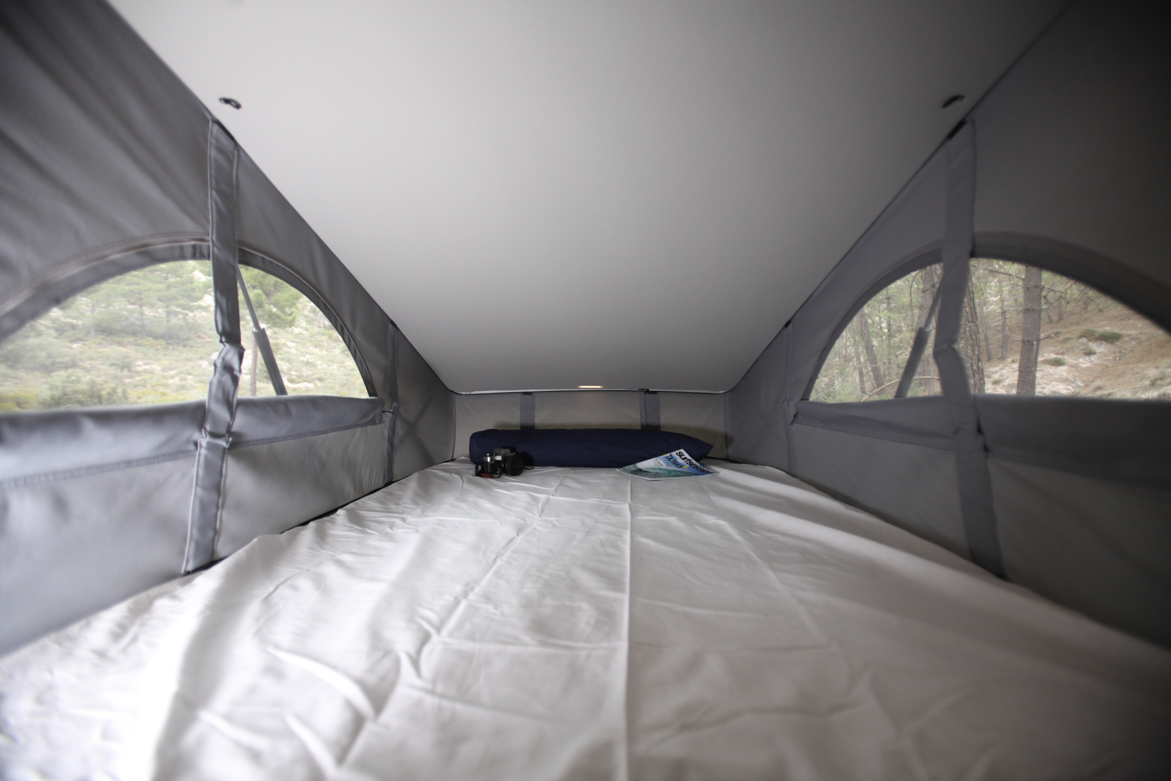 Inside an rv rental pop-top camper: white bed, grey walls, and arched windows showing forest views.. Volkswagen Transporter California Beach 2019