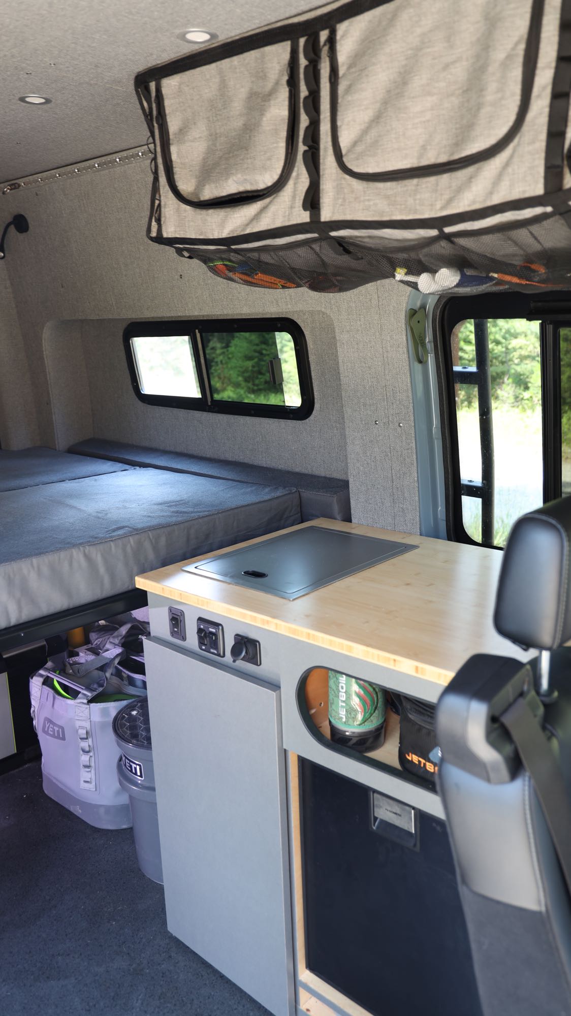 Modern RV rental interior showing a gray bed, light wood kitchen with sink, and practical overhead storage for adventure travel.. Mercedes-Benz Sprinter 2024