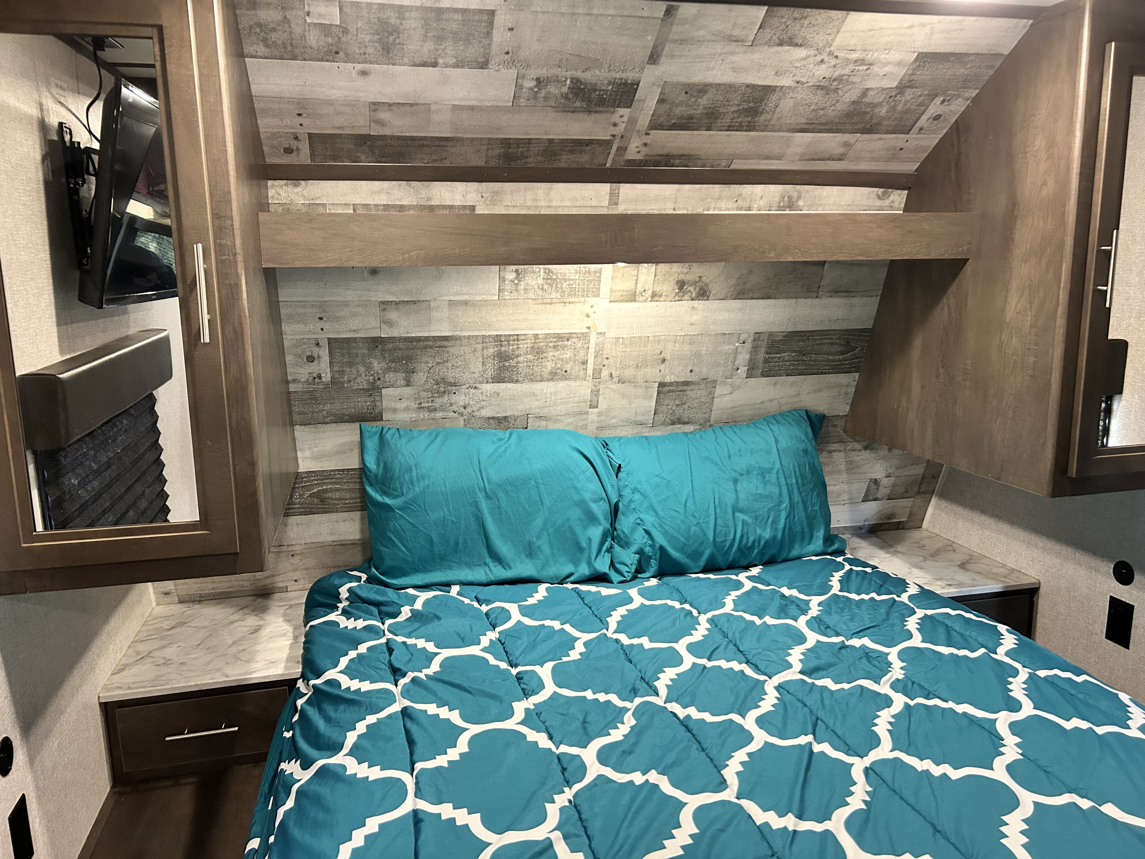 Experience comfort in this RV rental bedroom: cozy bed with teal linens, rustic wood paneling, TV, and smart storage.. Dutchmen Coleman Lantern 2021