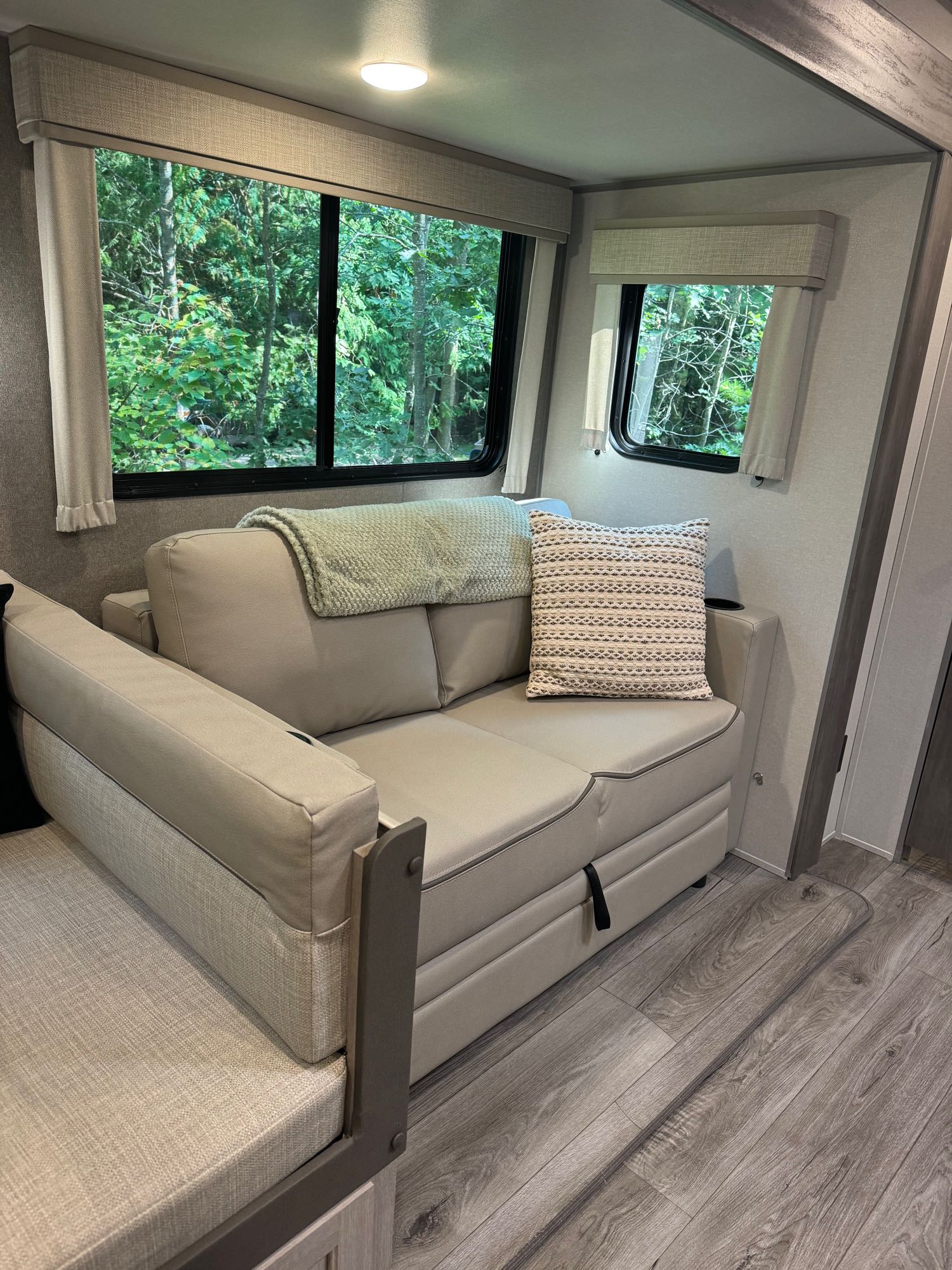 Comfortable RV rental living space featuring a light sofa, throw pillow, and blanket, with large windows overlooking a lush forest.. Coachmen Catalina 2024