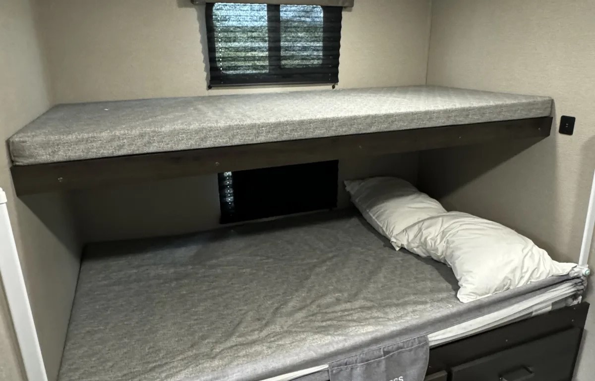 Inside an RV rental: a compact sleeping area with two bunk beds, grey mattresses, and a window. A white pillow rests on the bottom bunk.. Grand Design Transcend Xplor 2023