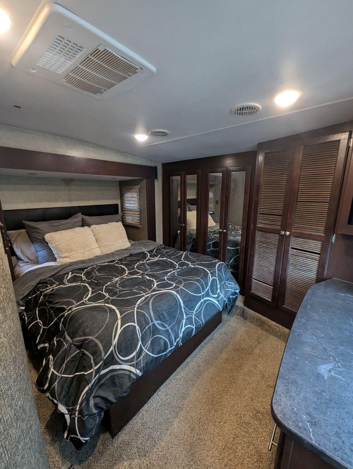 Comfortable RV rental bedroom featuring a patterned bed, overhead AC unit, and dark wood storage, perfect for your travels.. Heartland Road Warrior 2015