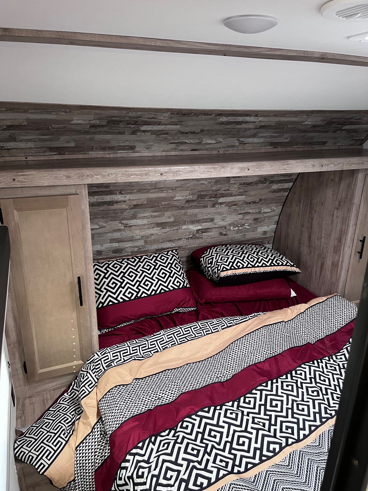 Cozy RV rental bedroom featuring a bed with maroon, black, and beige patterned bedding, built-in storage, and wood paneling.. Grand river 27BHS 2024