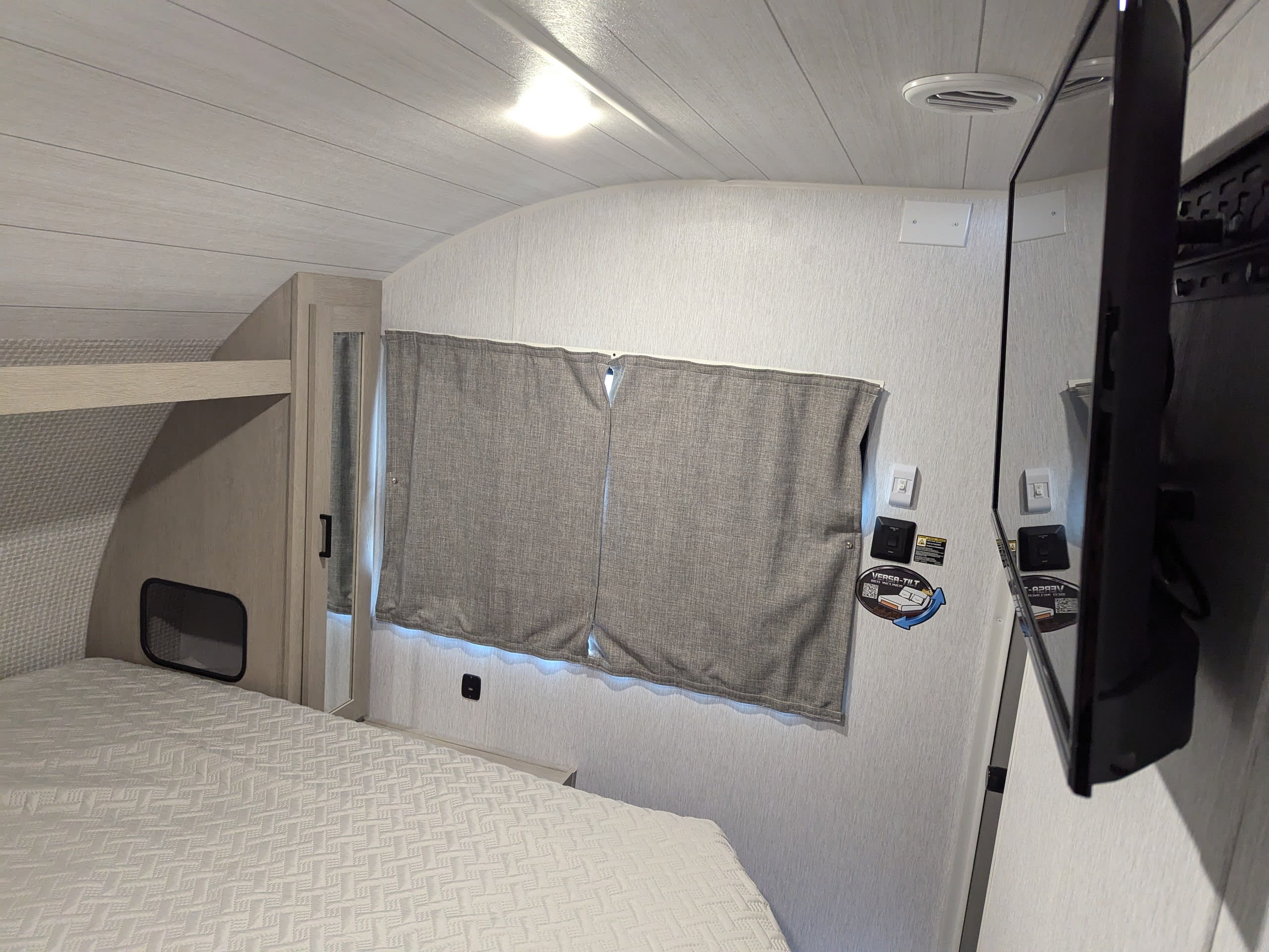 Cozy RV rental bedroom featuring a patterned bed, bunk, wall-mounted TV, and window curtains.. Forest River Wildwood X-Lite Platinum 2024