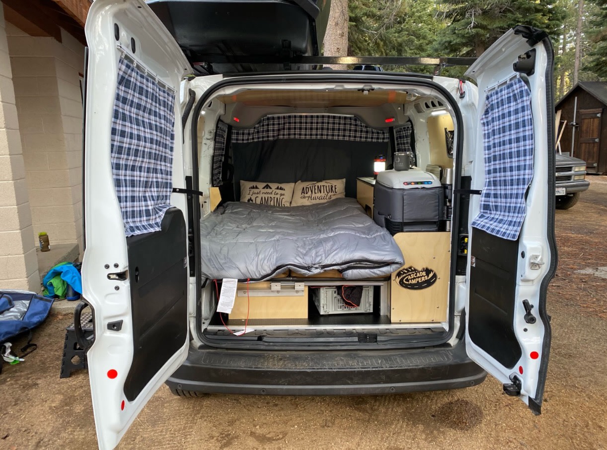 This RV rental camper van features a cozy bed with plaid curtains, fridge, and storage, ready for camping.. Cascade Campers Ram Promaster City 2020