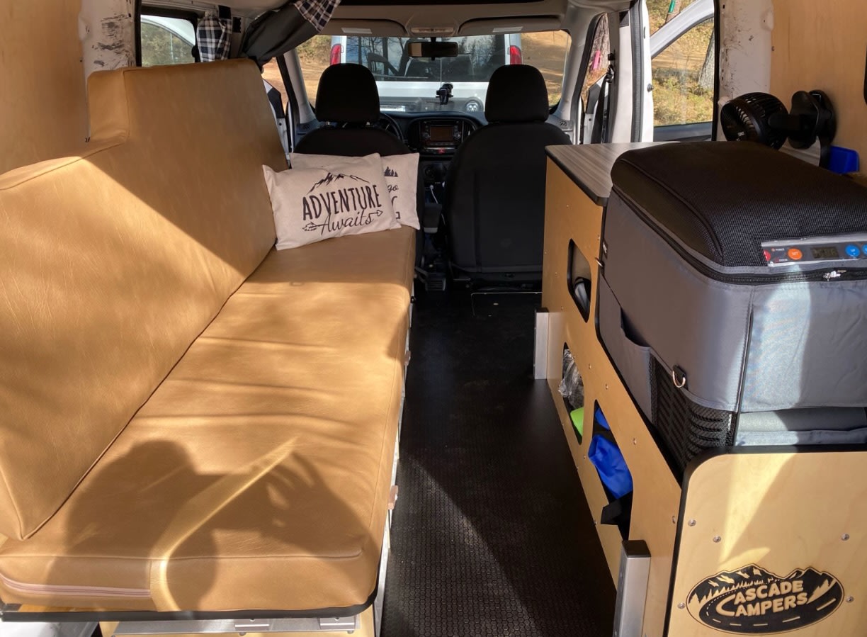 View inside an rv rental camper van. Features a long tan bench/bed on left, narrow aisle, and storage unit with portable fridge.. Cascade Campers Ram Promaster City 2020