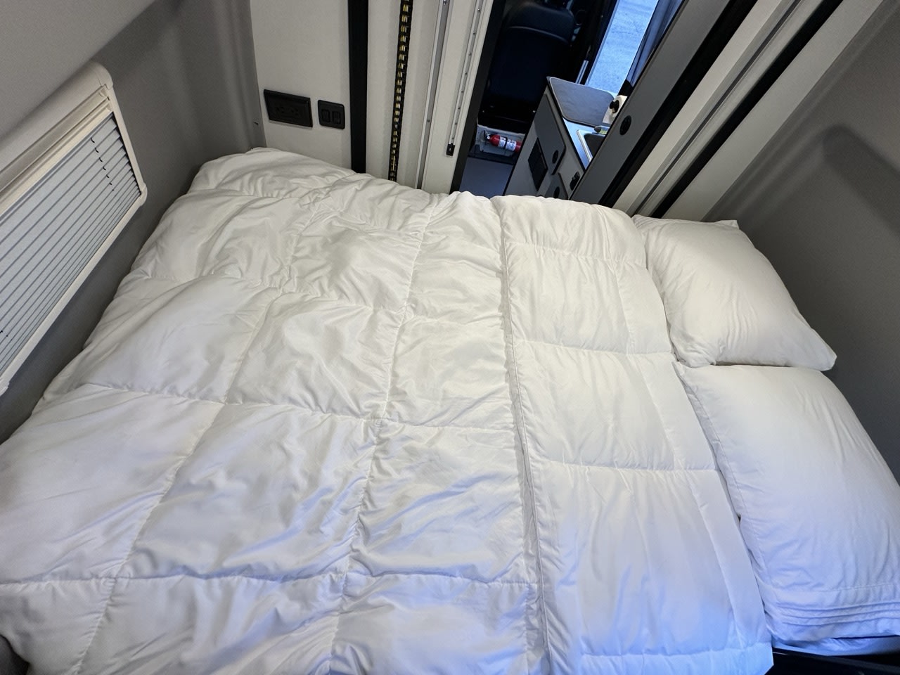 RV rental interior: cozy bed with white comforter and pillows, window, and compact kitchen area for comfortable travel.. Winnebago Revel 2023