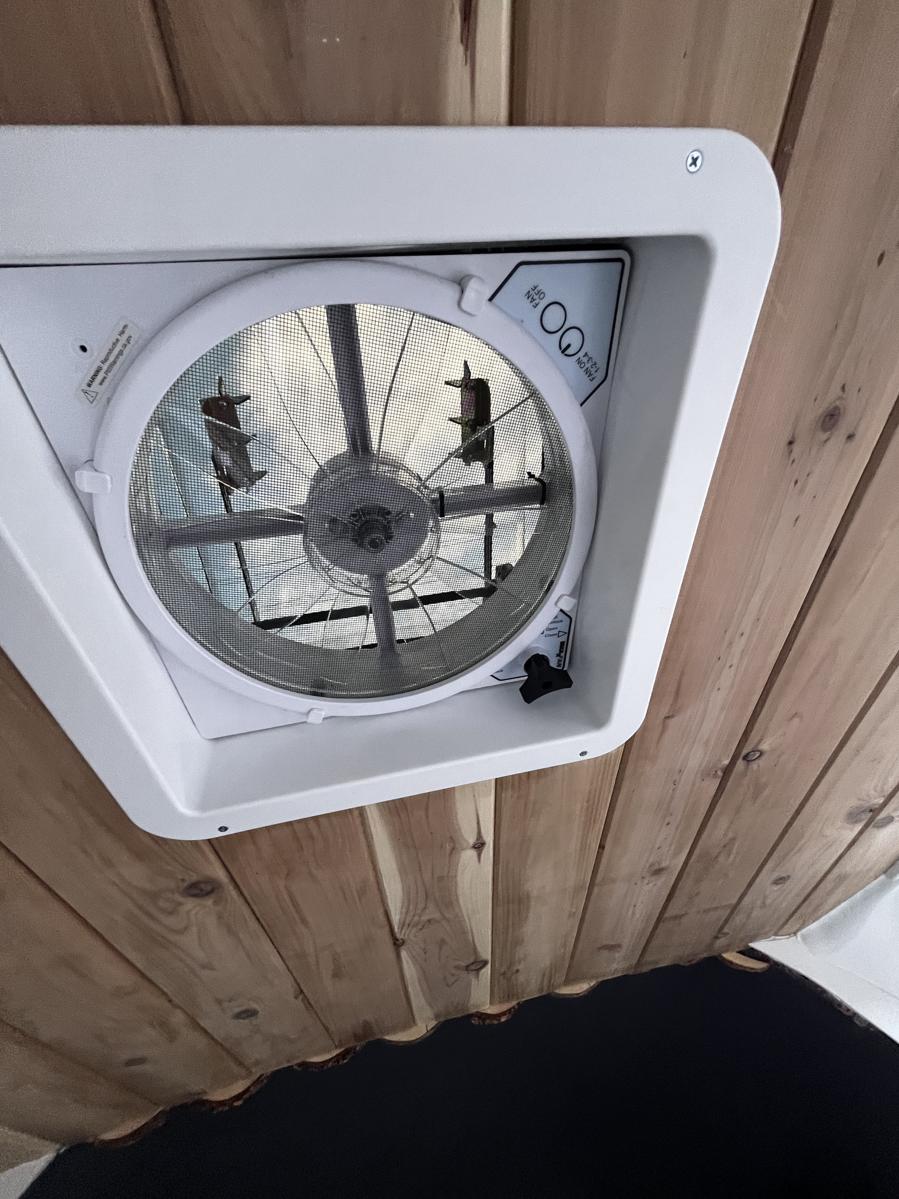 Overhead view of a white RV roof vent fan with a mesh screen against a rustic wood plank ceiling, common in an RV rental for airflow.. Mercedes-Benz Sprinter-Loaded 2022