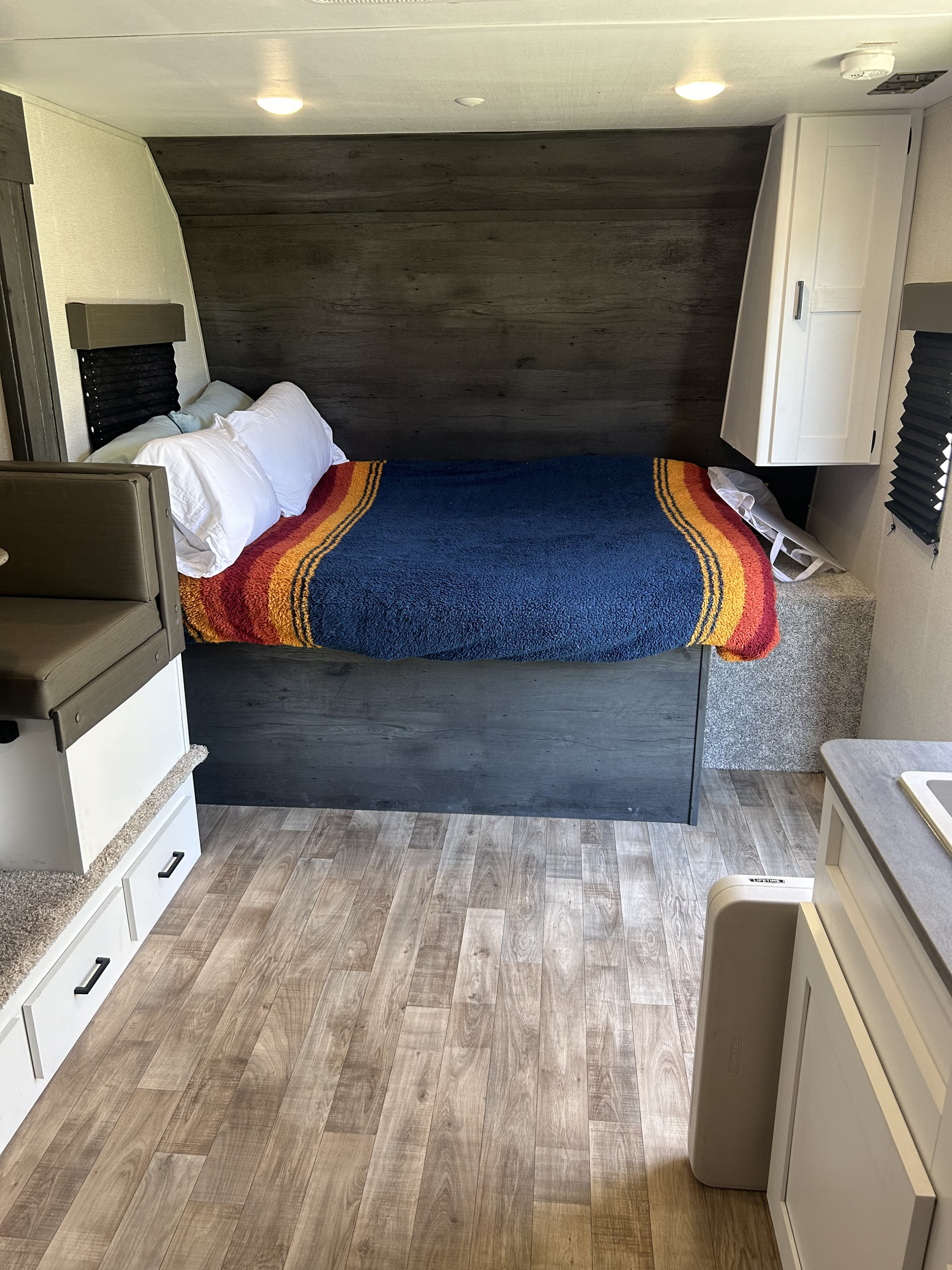 RV rental: cozy bedroom featuring dark wood-paneled walls, white pillows, colorful blanket on bed, and light wood flooring.. Keystone RV Hideout 2022