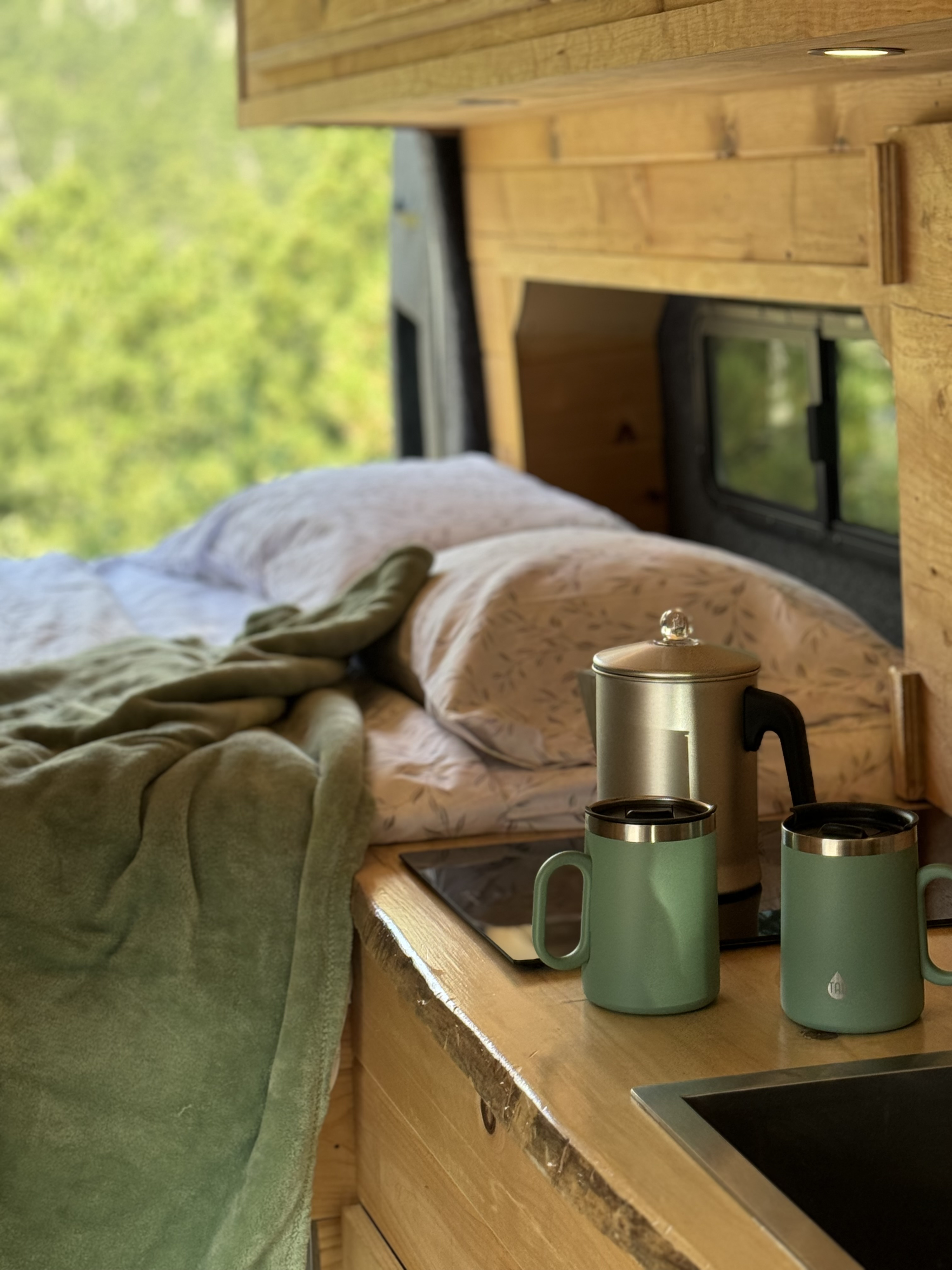 Cozy RV rental interior with a comfy bed, green blanket, and countertop featuring a coffee pot and two mugs, overlooking green trees.. Ford Transit 2019