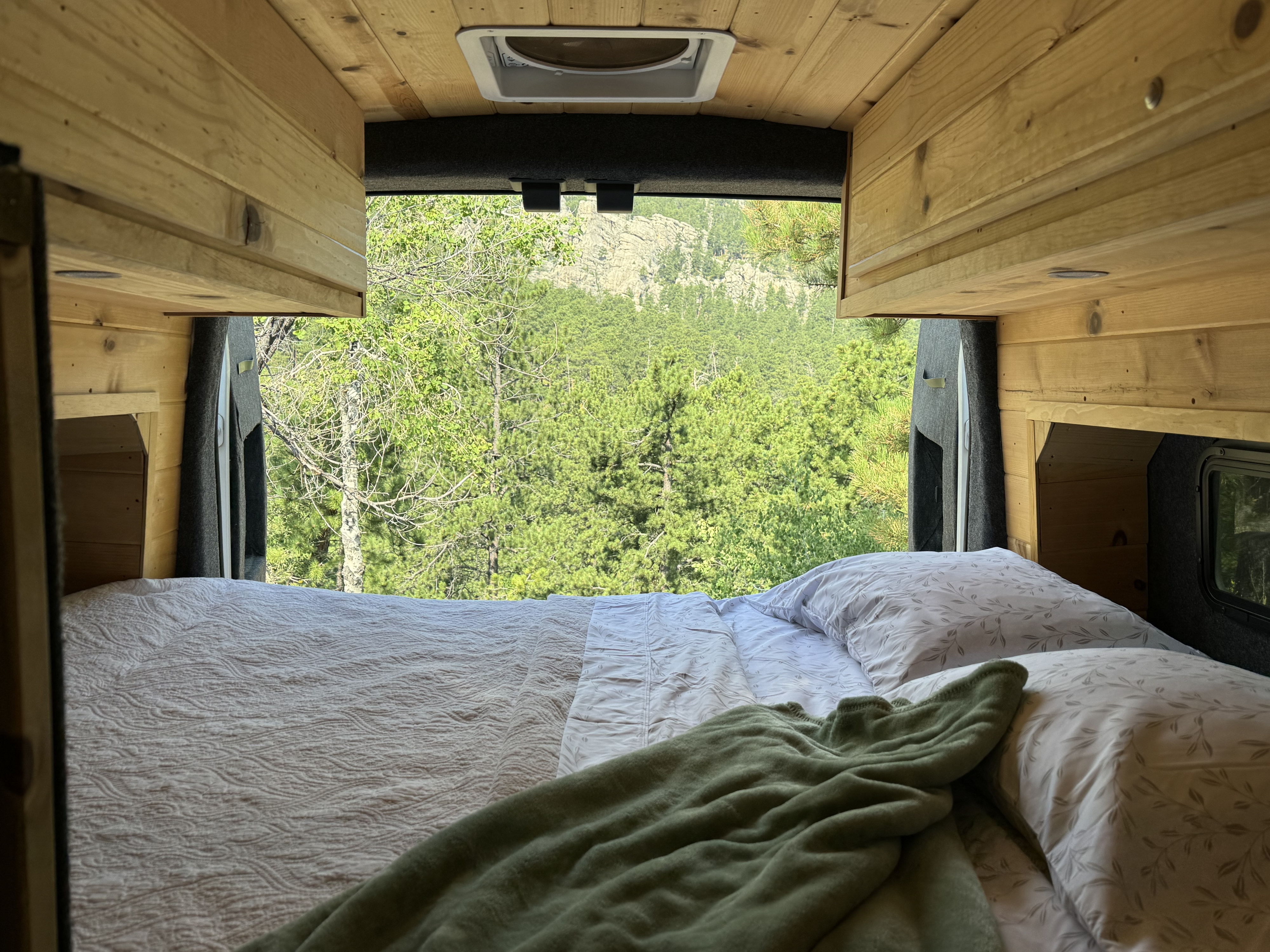 Inside a cozy RV rental: a comfortable bed offers panoramic views of a lush forest and mountains from the rear doors.. Ford Transit 2019