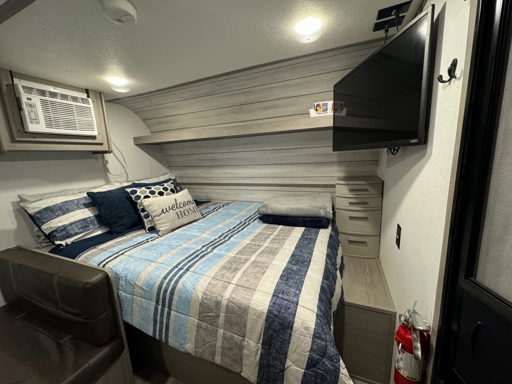 An RV rental bedroom setup: striped bed, mounted TV, AC unit, and a visible fire extinguisher for safety.. Coleman Lantern LT 17B 2024
