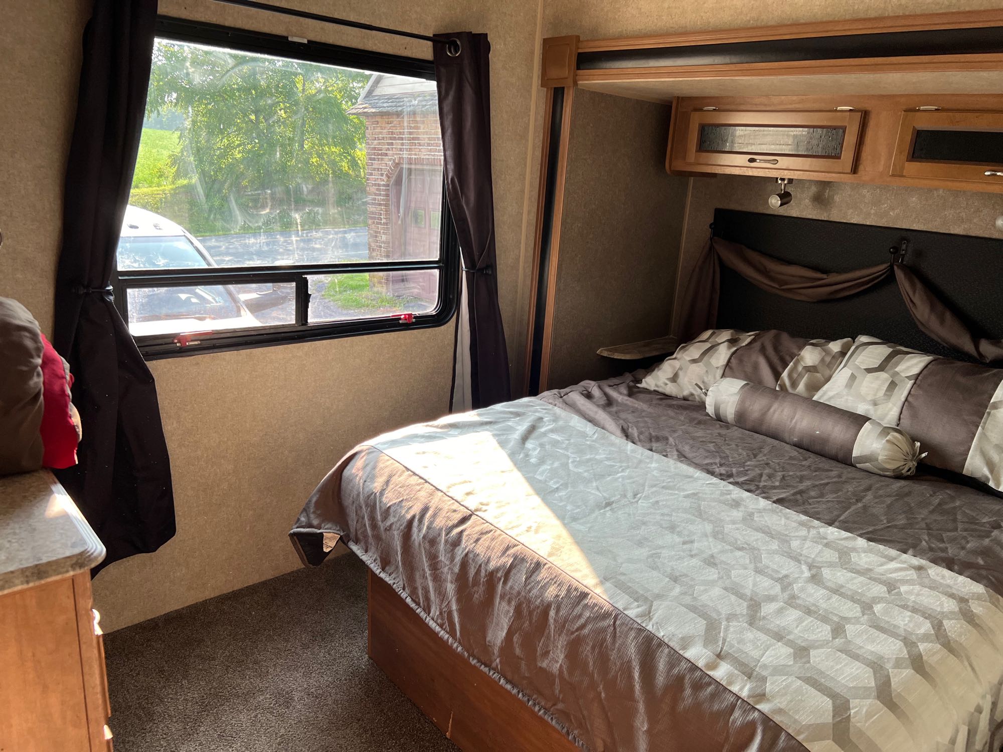 Comfortable RV rental bedroom featuring a bed with patterned linens, sunlit window view, and overhead storage. Ready for travel.. Coachmen Catalina 2018