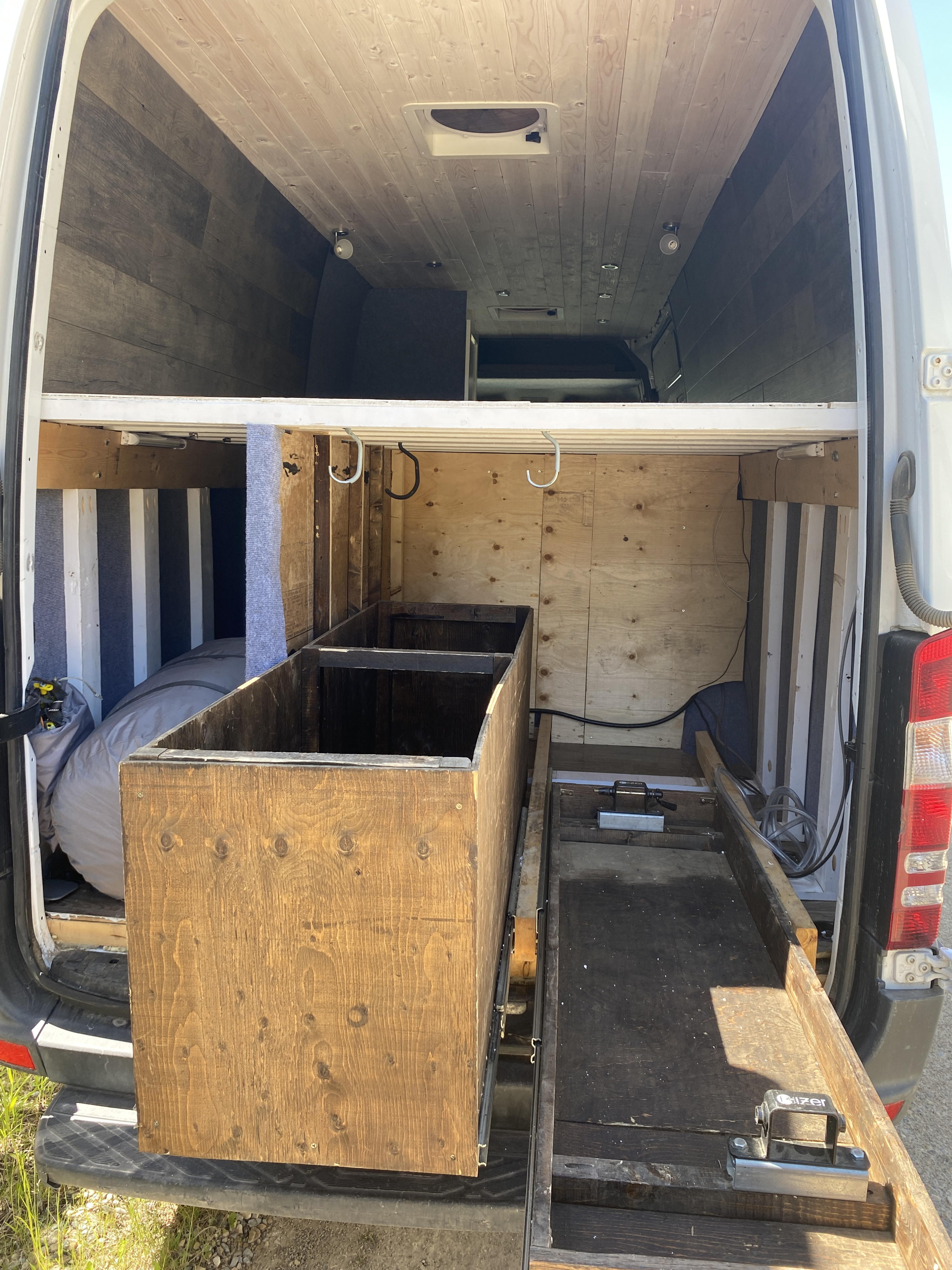 RV rental van's interior, featuring a wood-paneled ceiling, a bed, and a pull-out wooden storage box for organized travel.. Mercedes-Benz Sprinter 2013