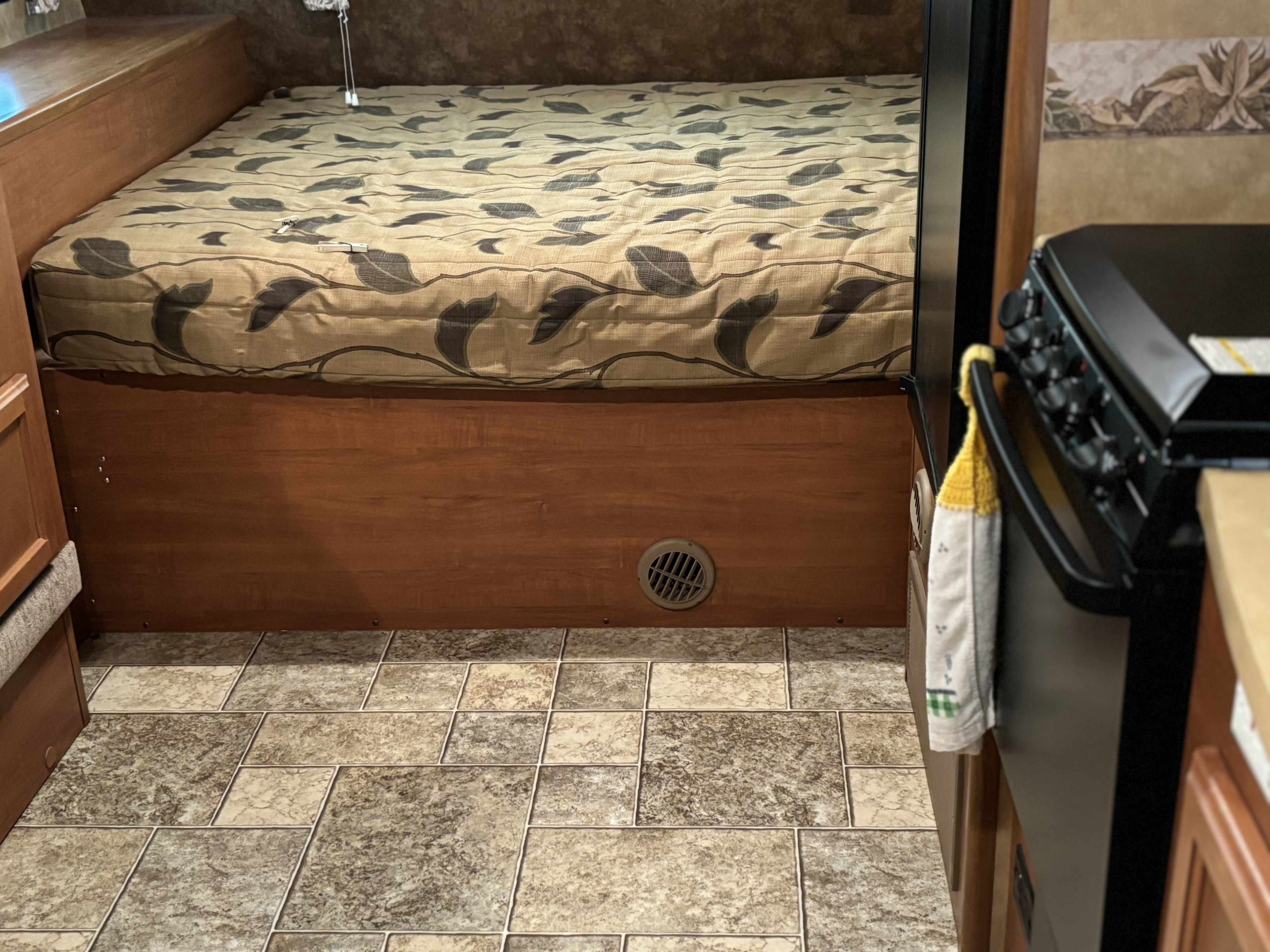 RV rental's sleeping area with a leafy-patterned mattress, wooden bed frame, and durable tiled floor, plus a glimpse of the kitchen.. Lance 1685 2012