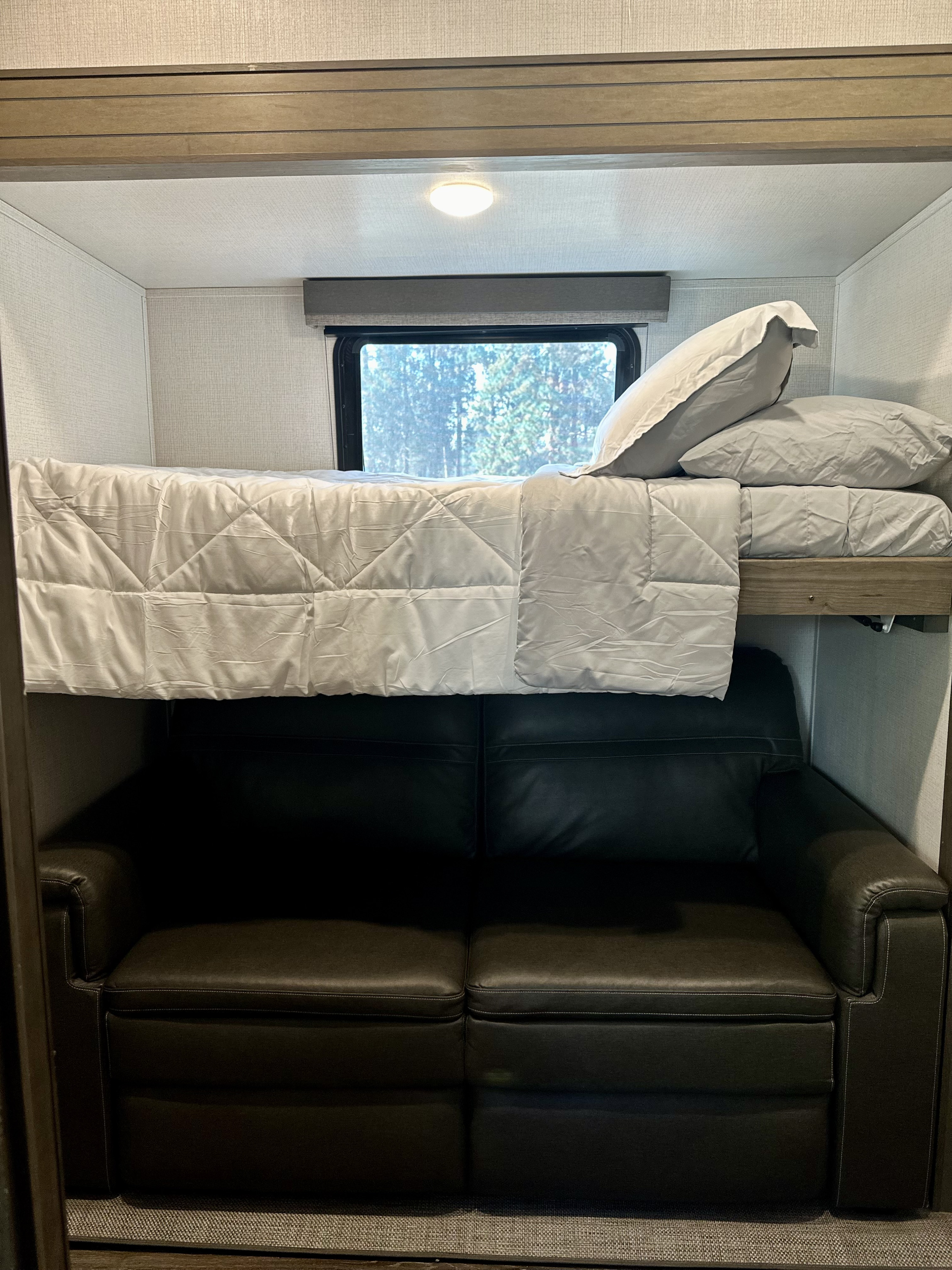 RV rental bunk bed with a soft quilt, pillows, and a window, positioned above a reclining leather sofa.. Keystone RV Cougar 2021