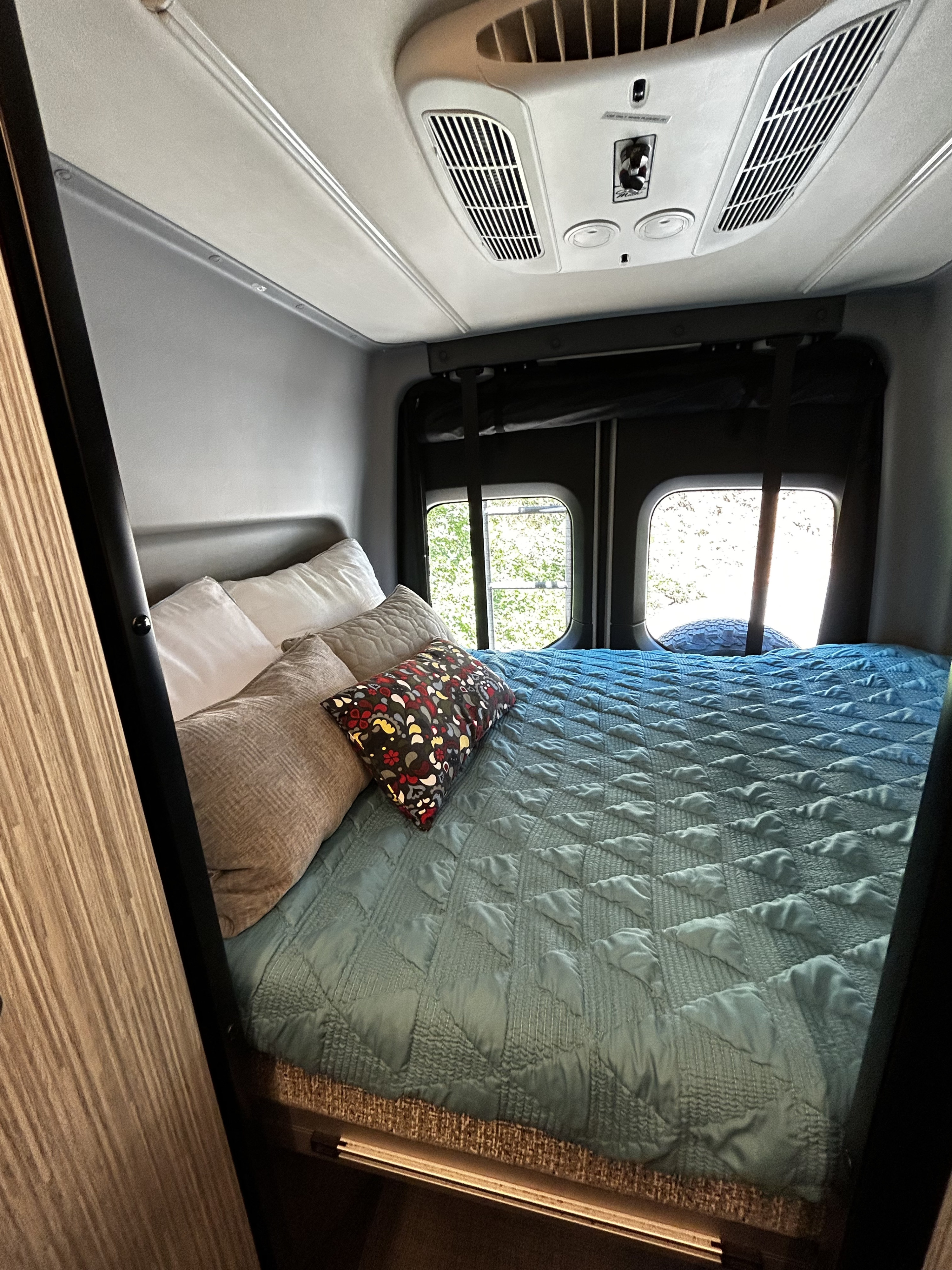 Cozy RV rental bedroom: blue quilted bed, pillows, rear windows, and overhead AC unit for comfortable travels.. Winnebago Revel 2020