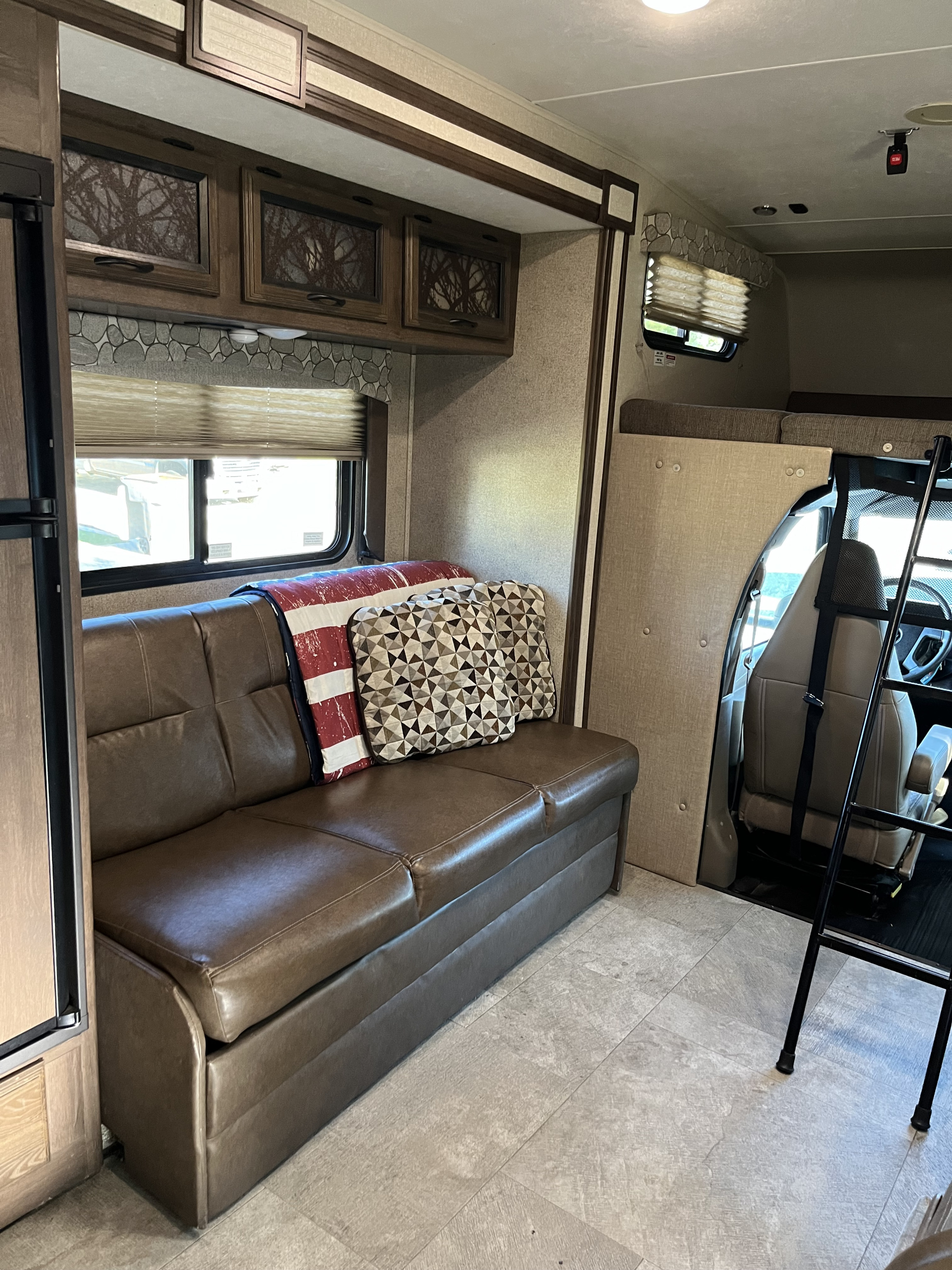 RV rental interior: living area with brown sofa, overhead cabinets, window, and front bunk access ladder.. Coachmen Freelander 2019