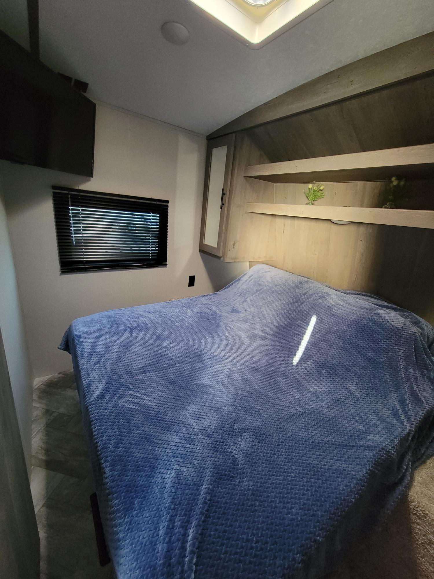 Cozy RV rental bedroom: blue blanket on bed, wall-mounted TV, window with blinds, and decorative shelving.. Keystone RV Springdale 2021