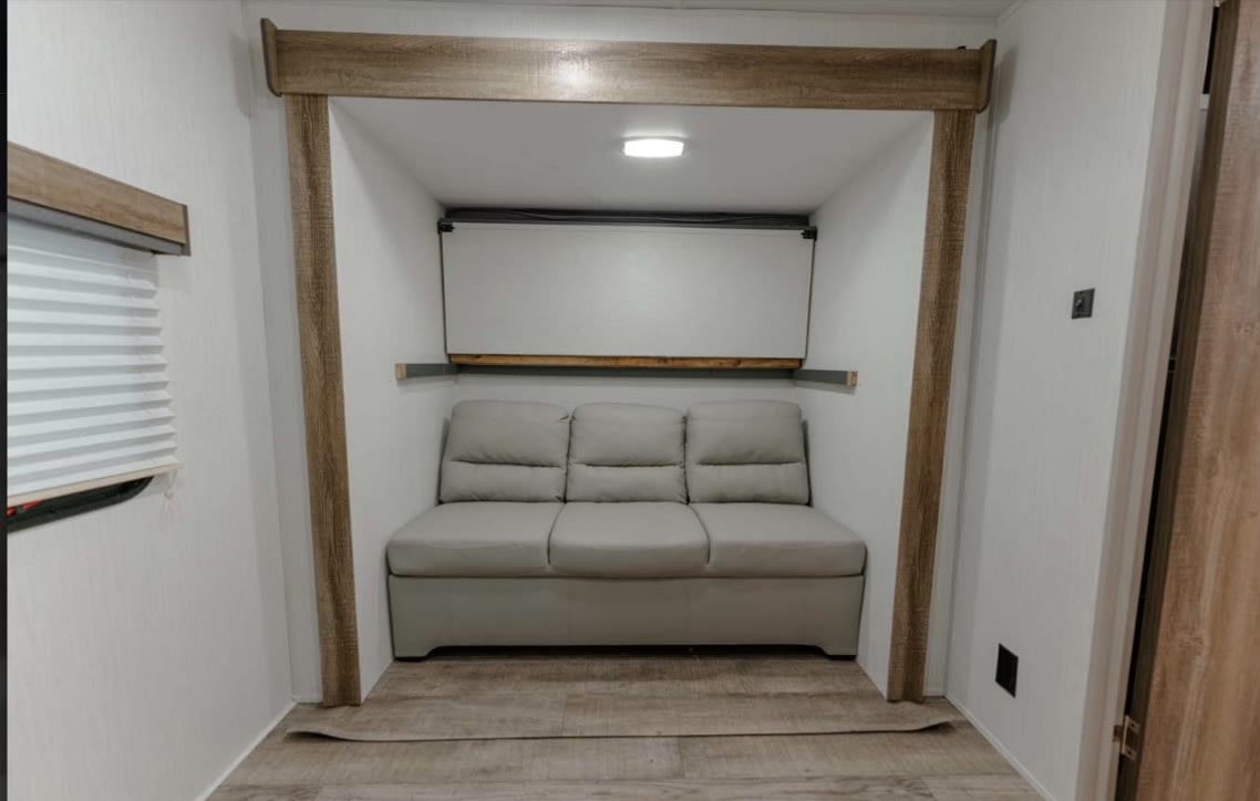 RV rental's cozy alcove: gray three-seat sofa, fold-down bed above, wood-look trim, and window blinds visible.. Heartland RVs Eddie Bauer BHS 34 2024