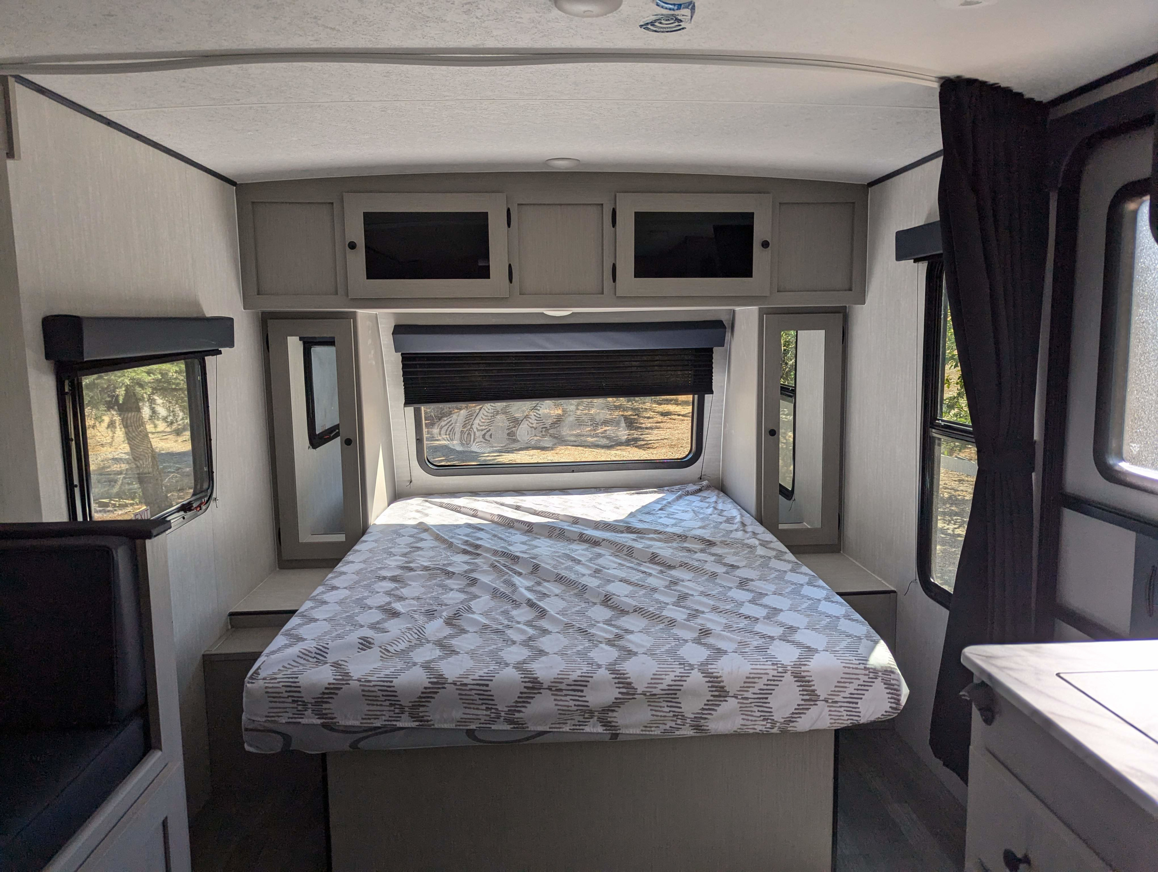 RV rental interior: a bright bedroom with a patterned bedspread, overhead cabinets, and windows showcasing nature outside.. Coachmen Apex Nano 2024