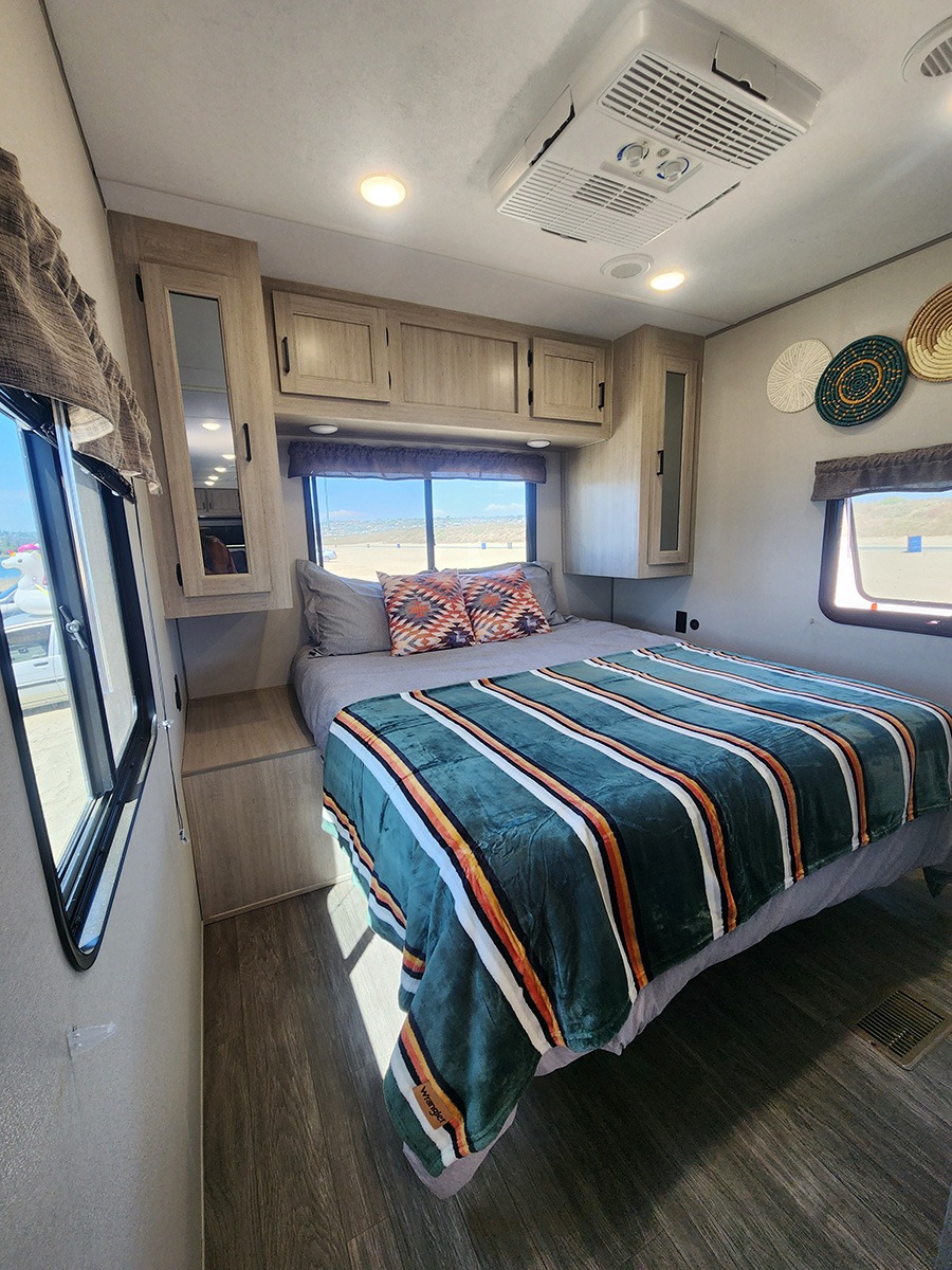 Inside a spacious RV rental bedroom featuring a comfy striped bed, overhead cabinets, AC unit, and windows with outdoor views.. Forest River Aurora Aurora 2024