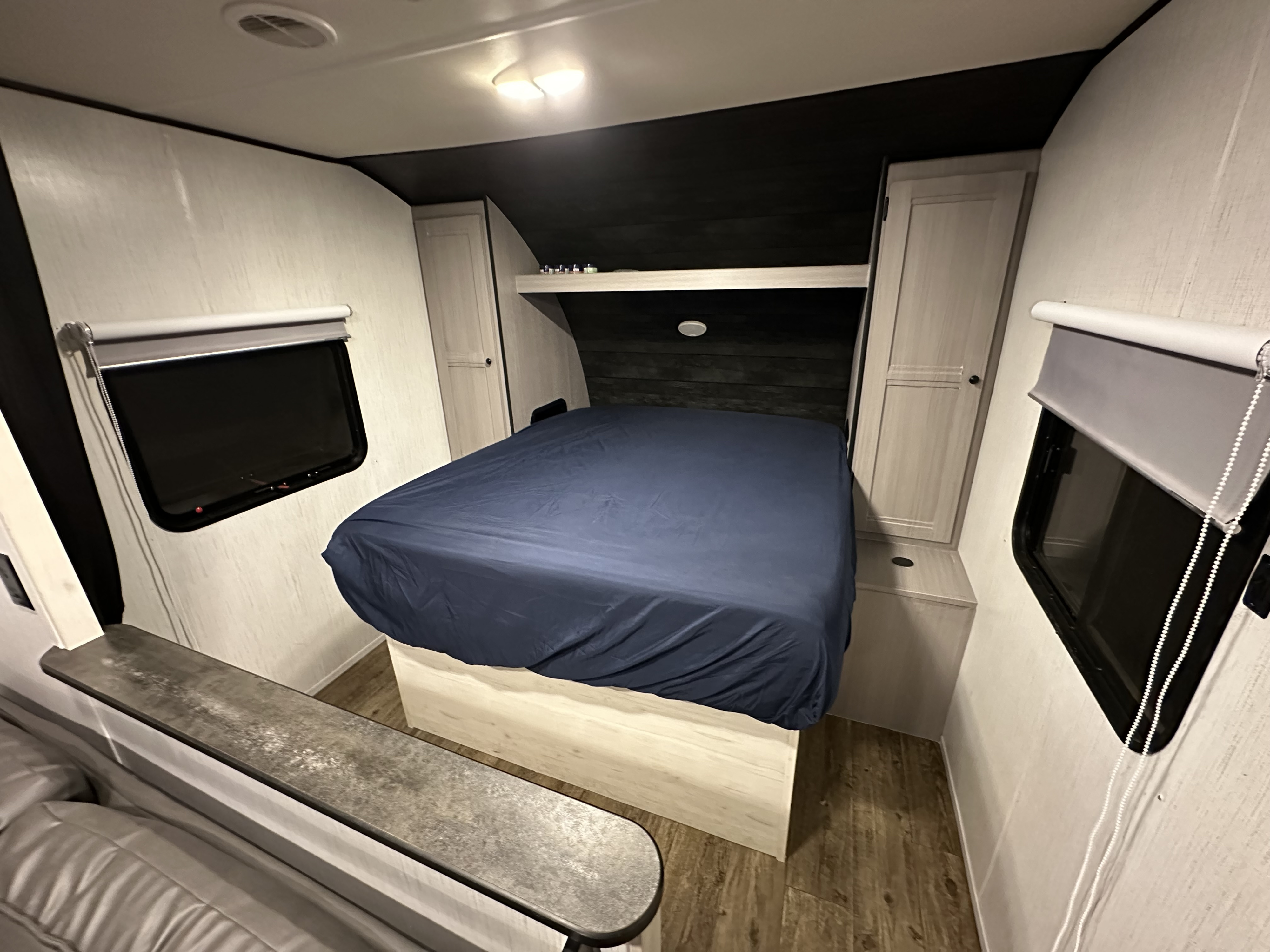 Modern RV rental bedroom featuring a bed with a blue cover, side storage, windows with shades, and light wood-look flooring.. Heartland RVs Trail Runner 2022